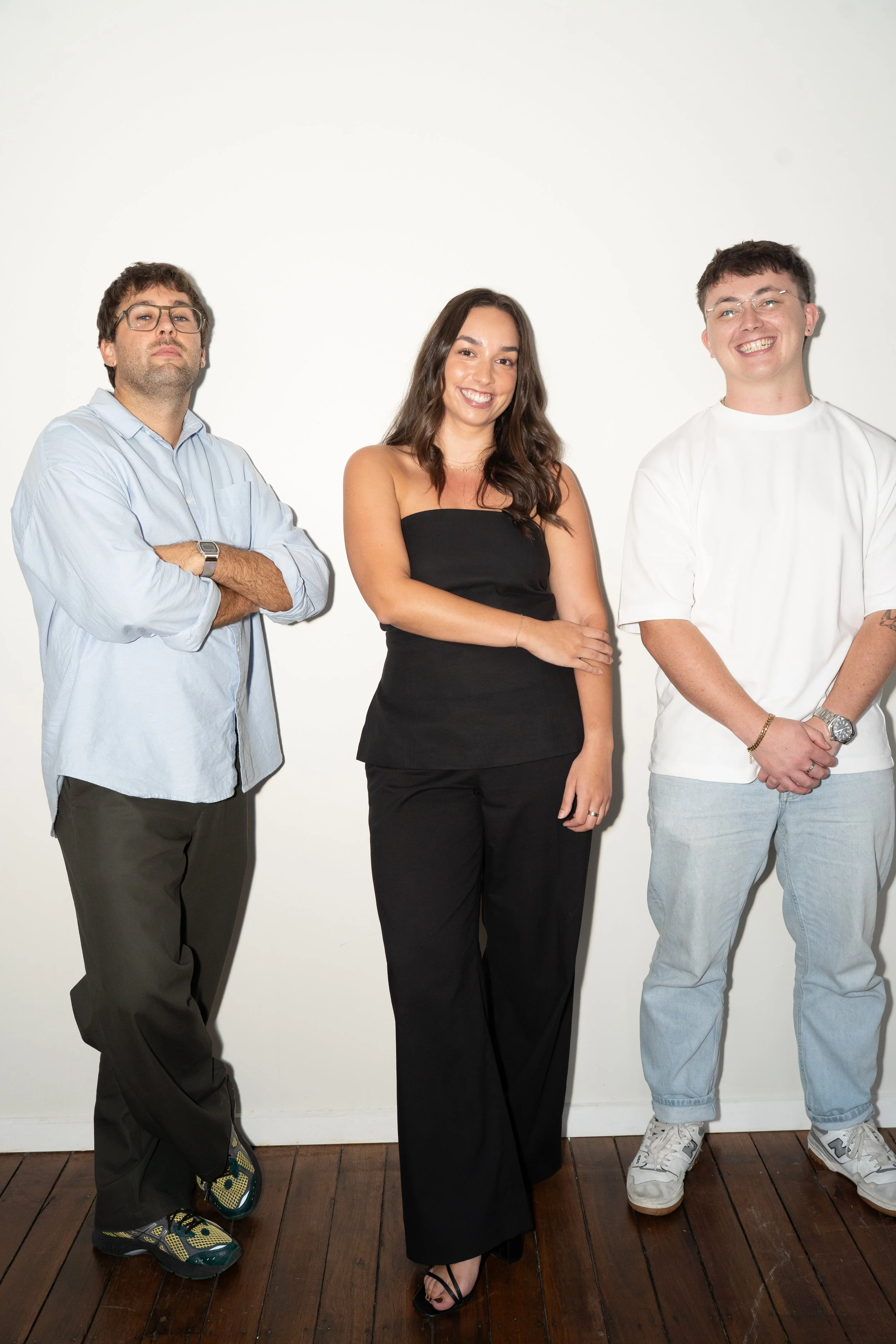 Three young adults standing against a white wall. The person on the left is a man with glasses, arms crossed, wearing casual clothes and sneakers. The person in the middle is a woman with long dark hair, smiling, wearing a black strapless top and black pants. The person on the right is a man with glasses, smiling broadly, wearing a white t-shirt and light blue jeans.