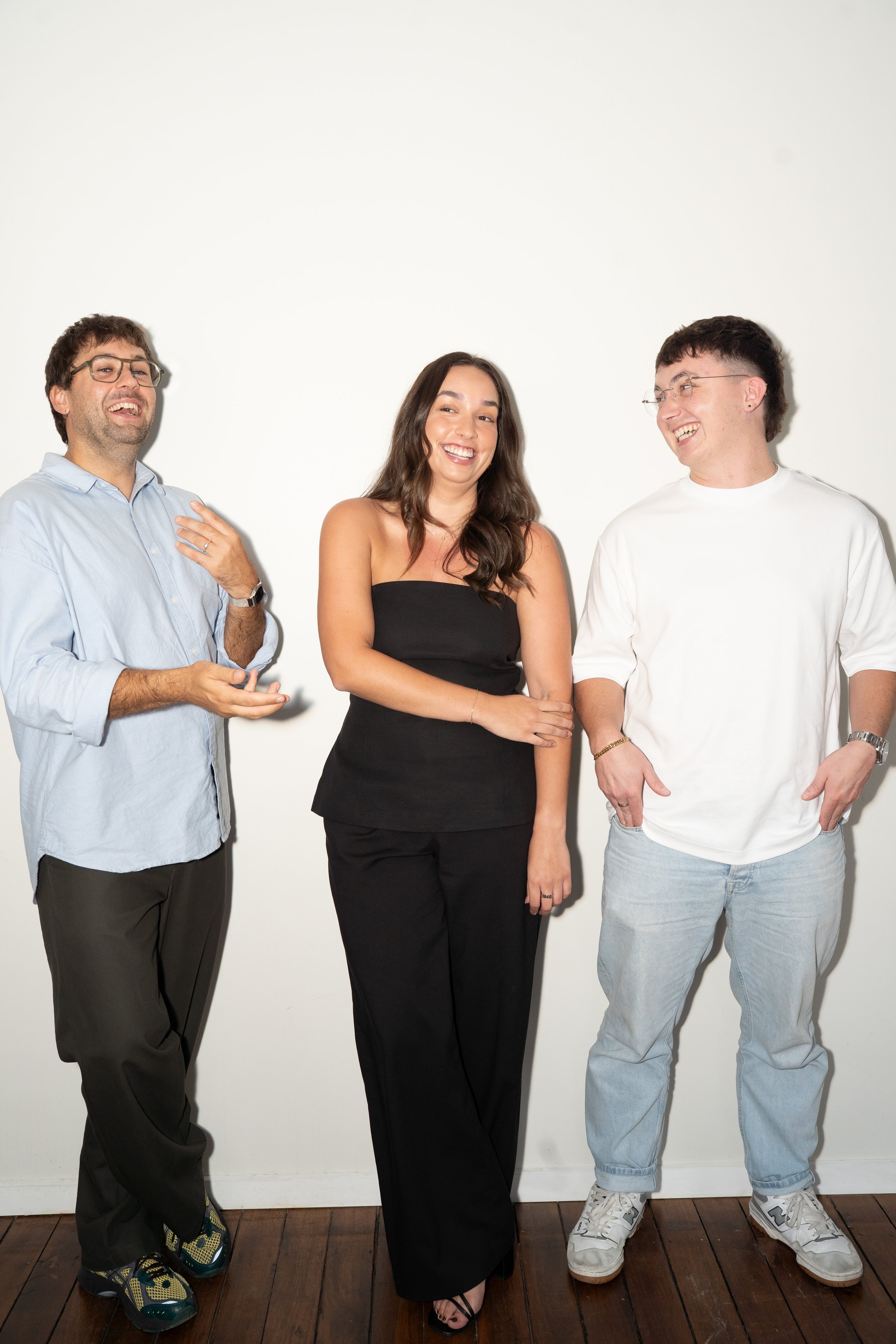 Three people standing in front of a plain white wall, smiling and engaged in conversation. The person on the left is a man with curly brown hair, glasses, wearing a light blue shirt and dark pants. The person in the middle is a woman with long dark hair, wearing a black strapless top and black pants. The person on the right is a man with short dark hair, glasses, wearing a white oversized T-shirt and light blue jeans.