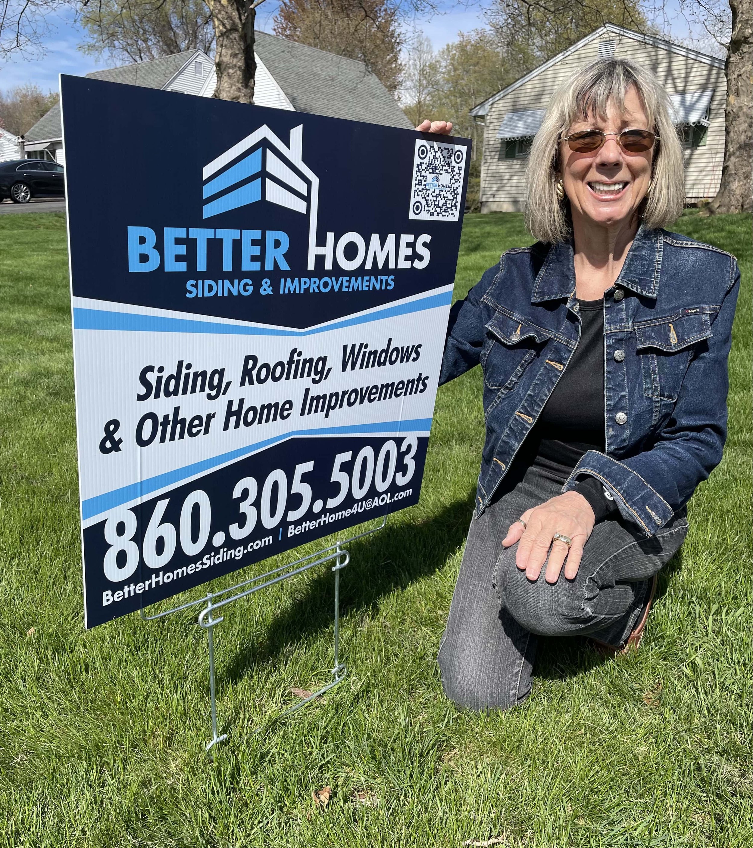 A woman kneeling on grass holding a sign for Better Homes siding and home improvements, with a house and trees in the background.