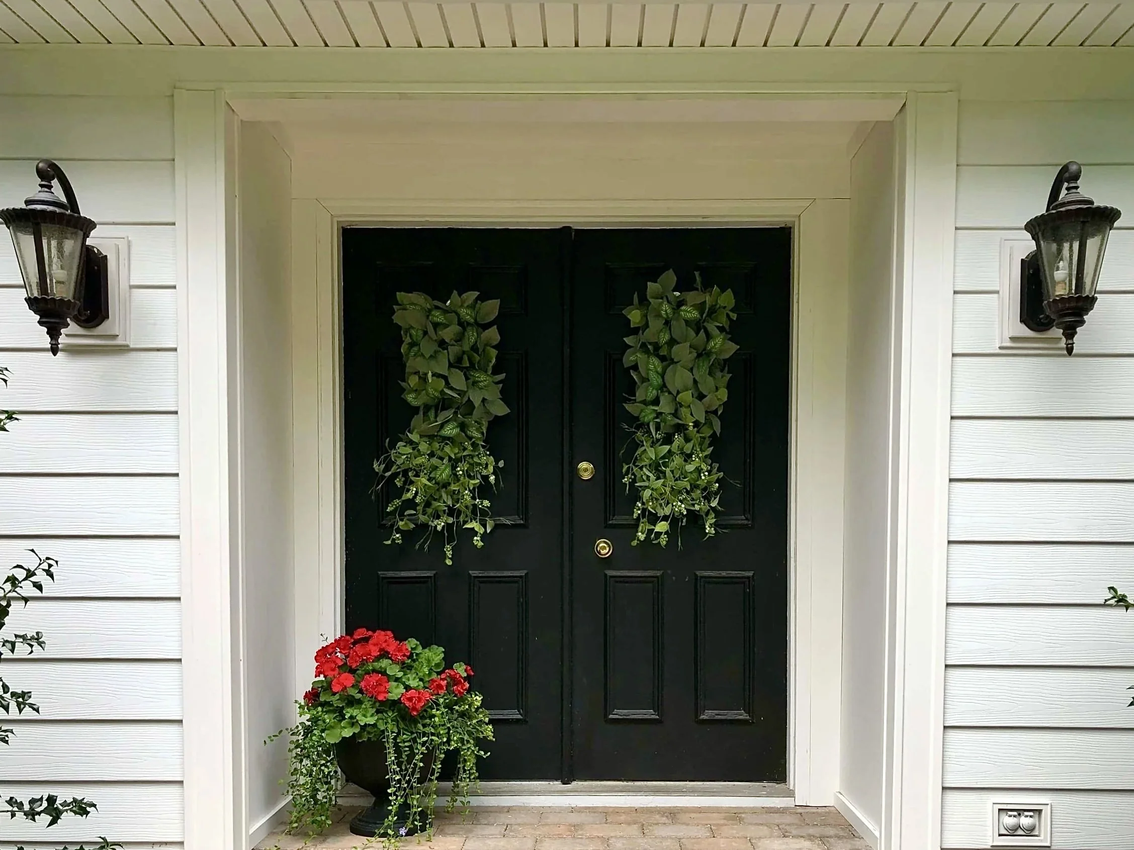 A black front door decorated with green leafy plants on both sides, flanked by two black wall-mounted outdoor lanterns, one on each side.