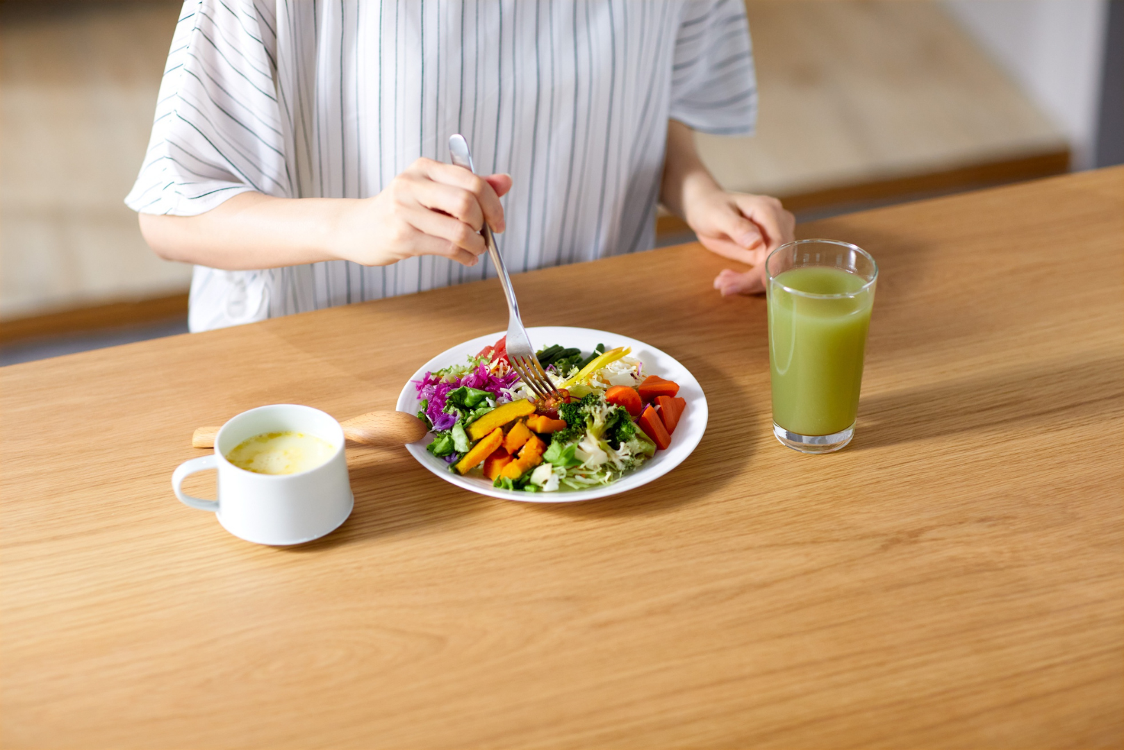 Woman eating a colorful plate of vegetables with soup and a green drink on a wooden table, illustrating a healthy, balanced meal and vibrant nutrition habits.