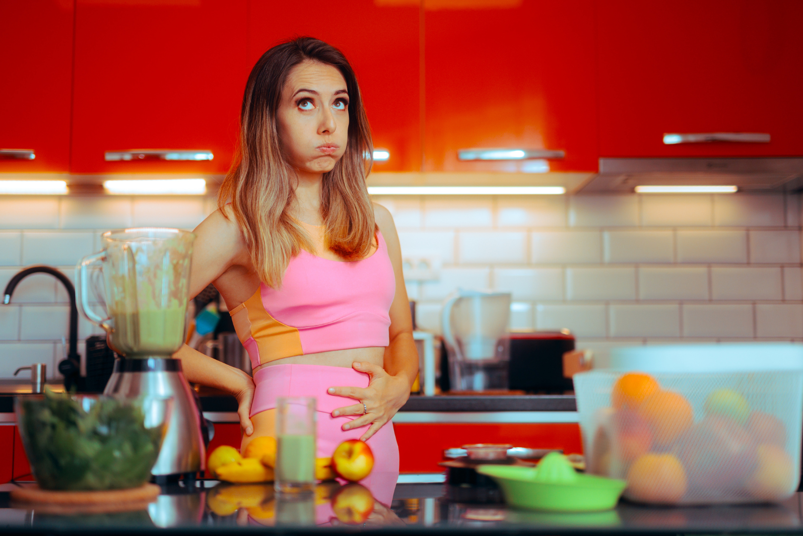 Woman in workout clothes holding her bloated stomach in a kitchen with a green smoothie, looking frustrated after detoxing but still feeling uncomfortable