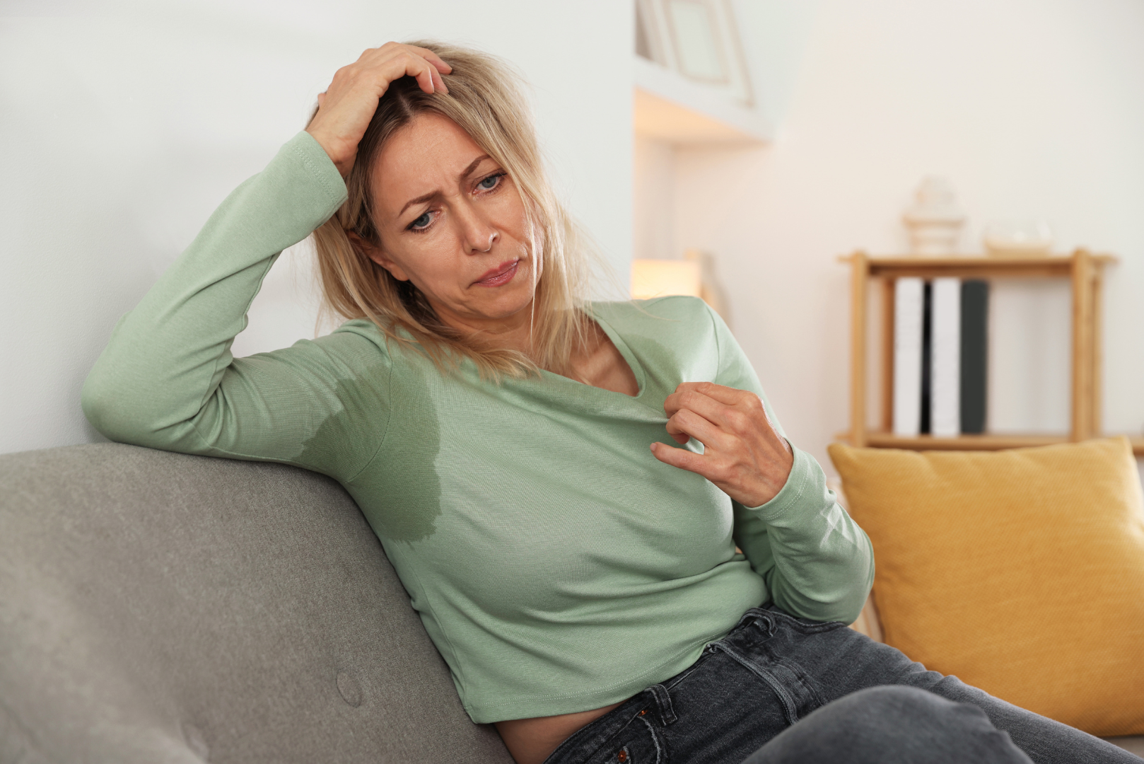 A middle-aged woman sitting on a couch, looking exhausted and pulling at her sweat-soaked shirt after a hot flash.