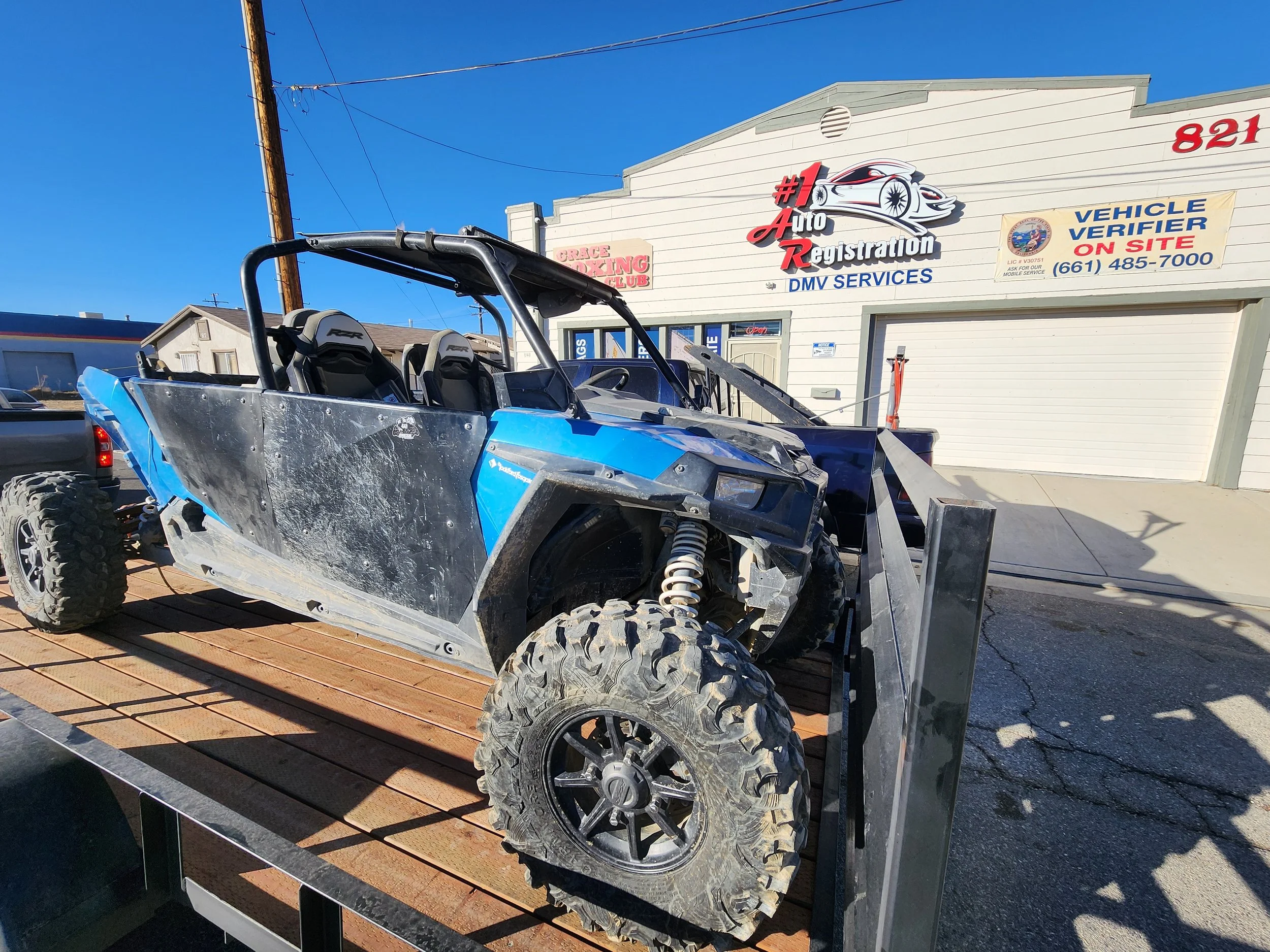 Off-road all-terrain vehicle, or UTV, with damaged body panels and dirt on tires, on a flatbed trailer in front of a building with a sign for auto registration services.