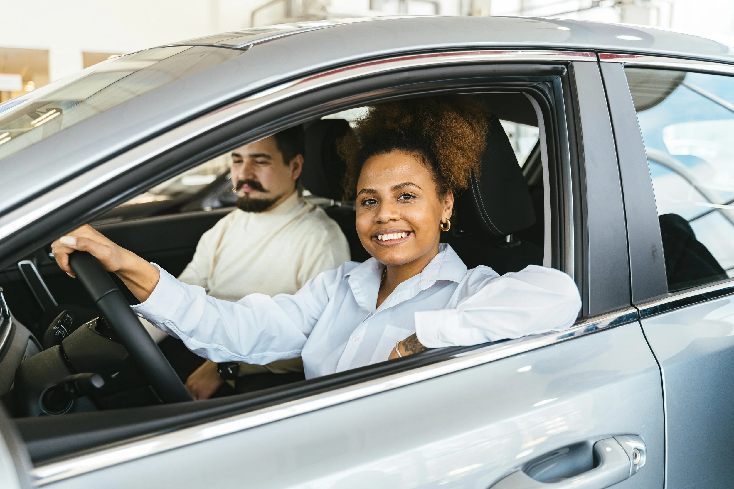 A woman with curly hair smiling and sitting in the driver's seat of a car, with a man with a mustache and beard sitting in the passenger seat, both inside a modern car showroom.