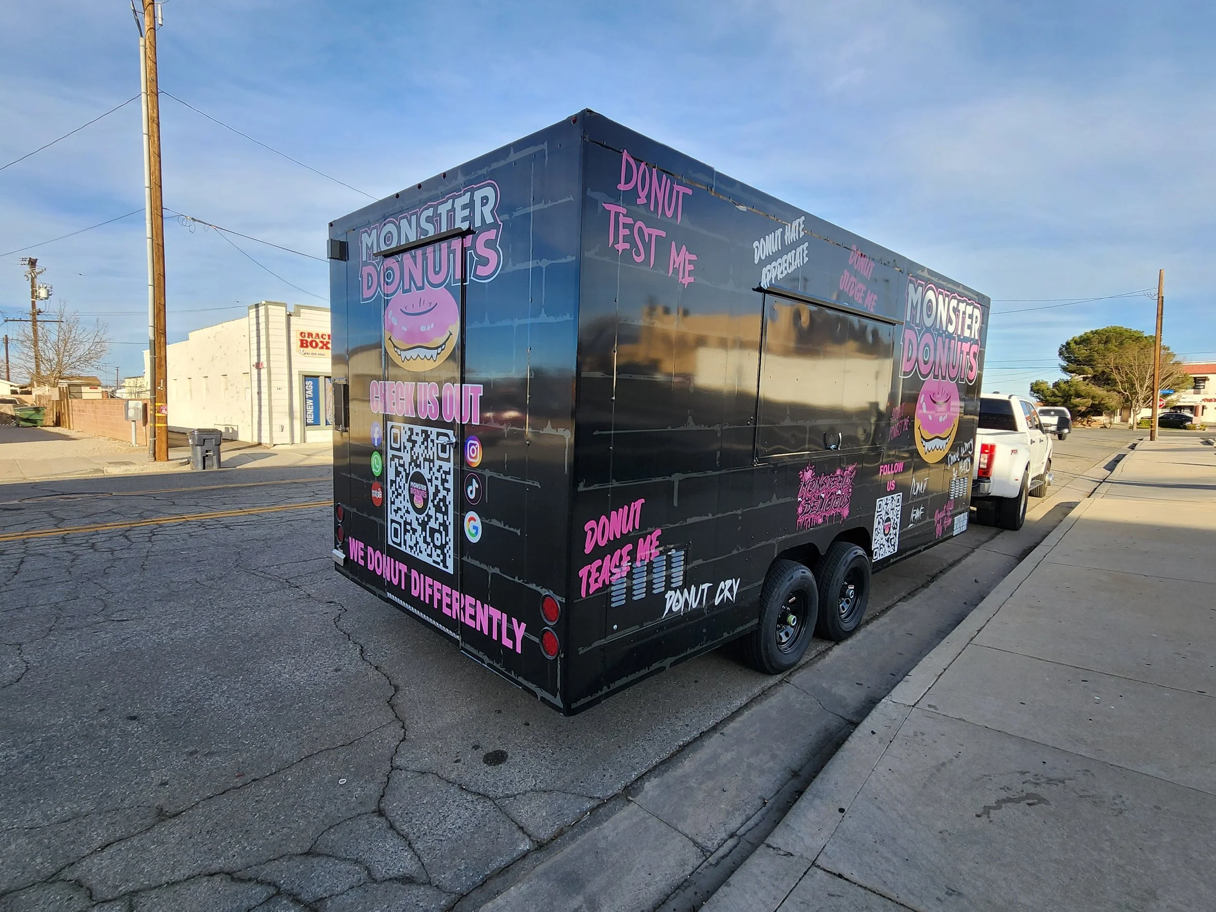 A black food trailer decorated with pink and white text and graphics advertising Monster Donuts. It features a cartoon donut logo, QR codes, and social media icons.