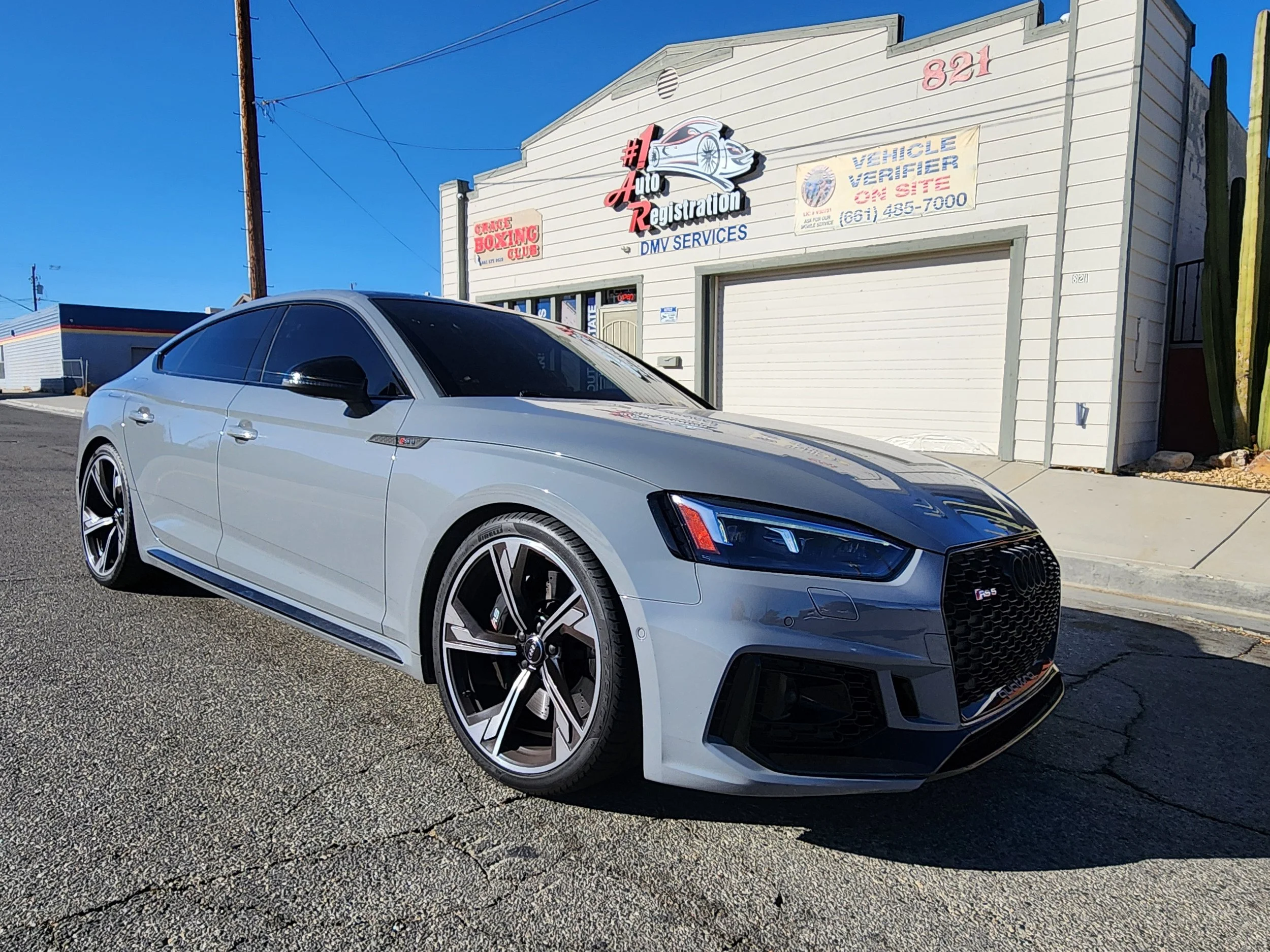 Silver Audi RS5 parked outside a building with signs for vehicle registration and DMV services.