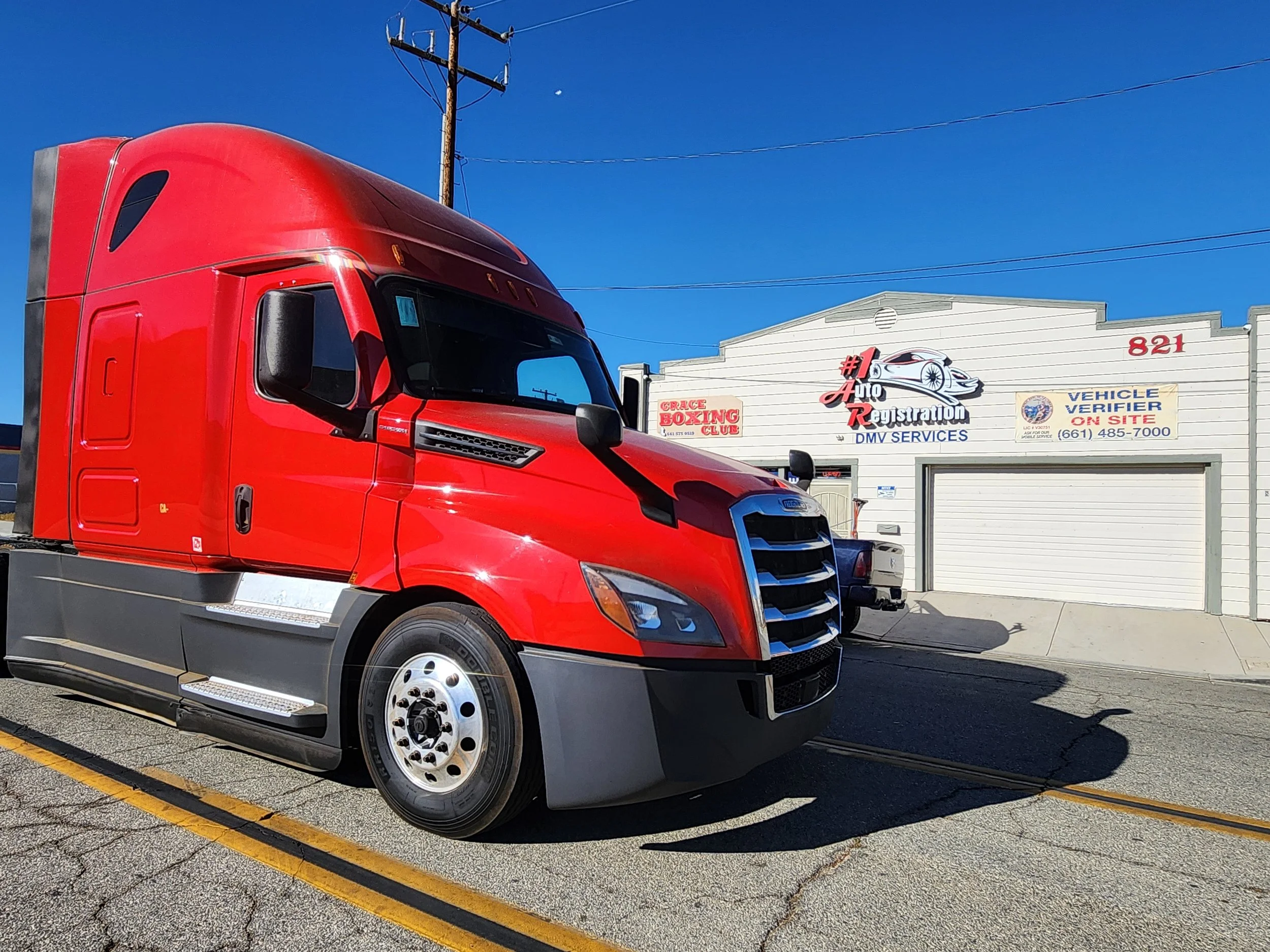 A red semi-truck parked on a street in front of a building with signs for auto registration and DMV services under a clear blue sky.