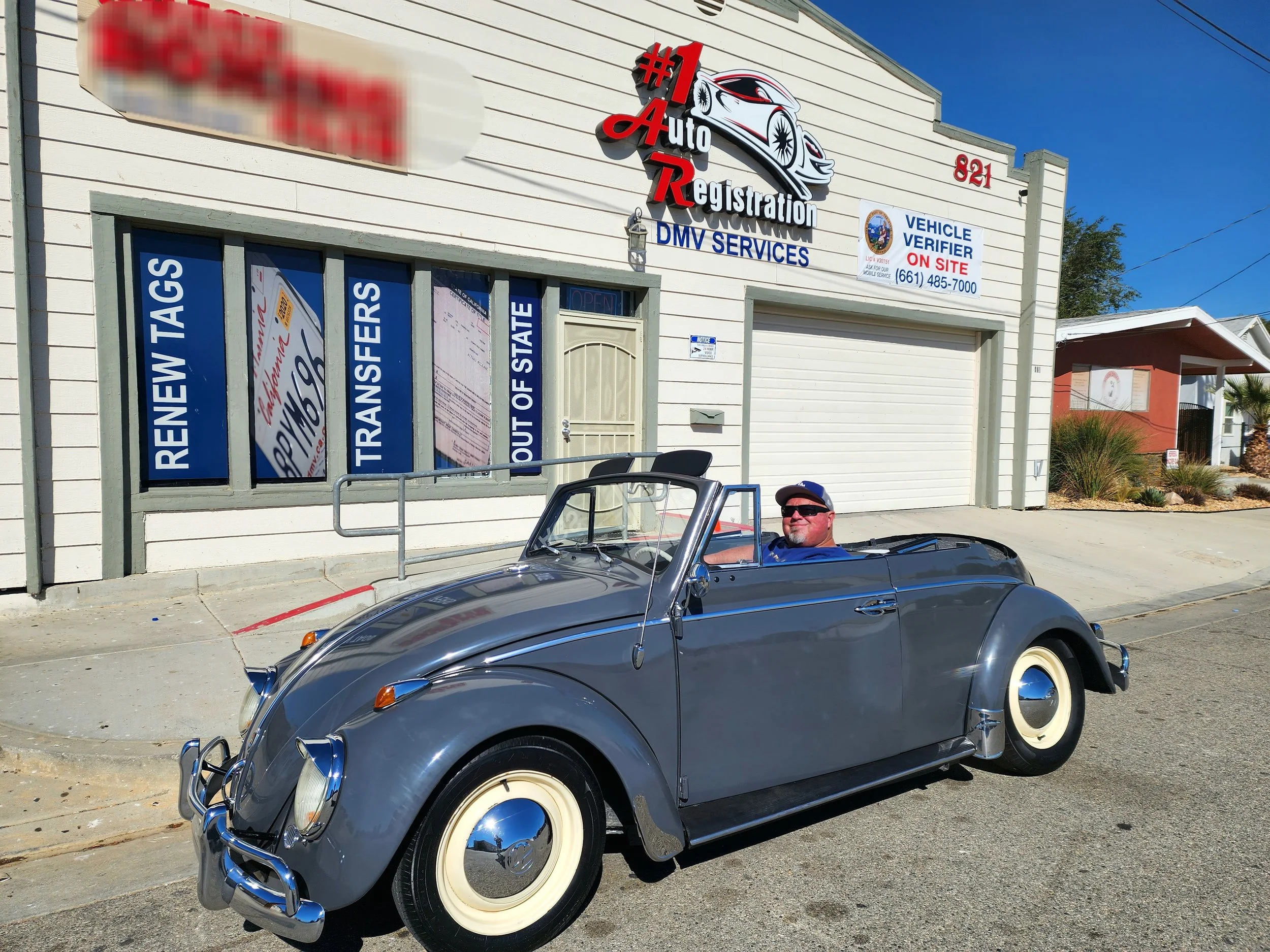 A man sitting in a vintage gray convertible car parked in front of an auto registration and DMV services business. The building has signage promoting vehicle tags, transfers, and out-of-state registrations.