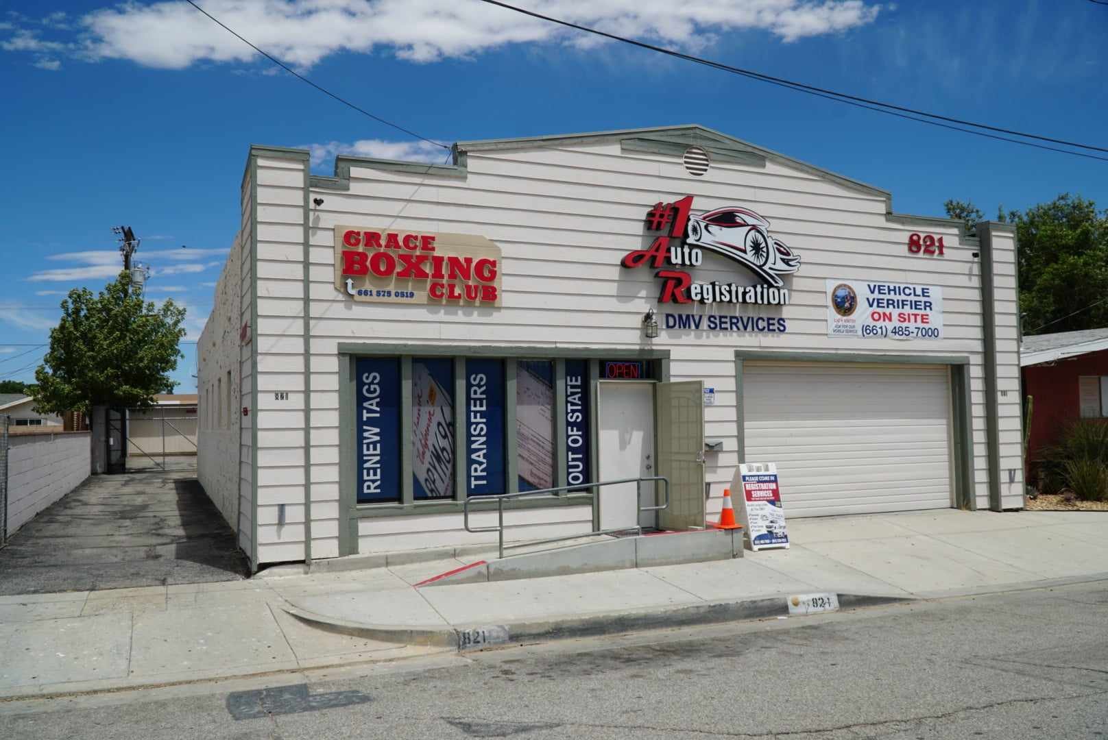 Exterior view of a white building with signs for auto registration, DMV services, vehicle verifier, and a boxing club. The building has a parking garage with a roller door and a sidewalk with an orange traffic cone and sign. The sky is mostly clear with some clouds.