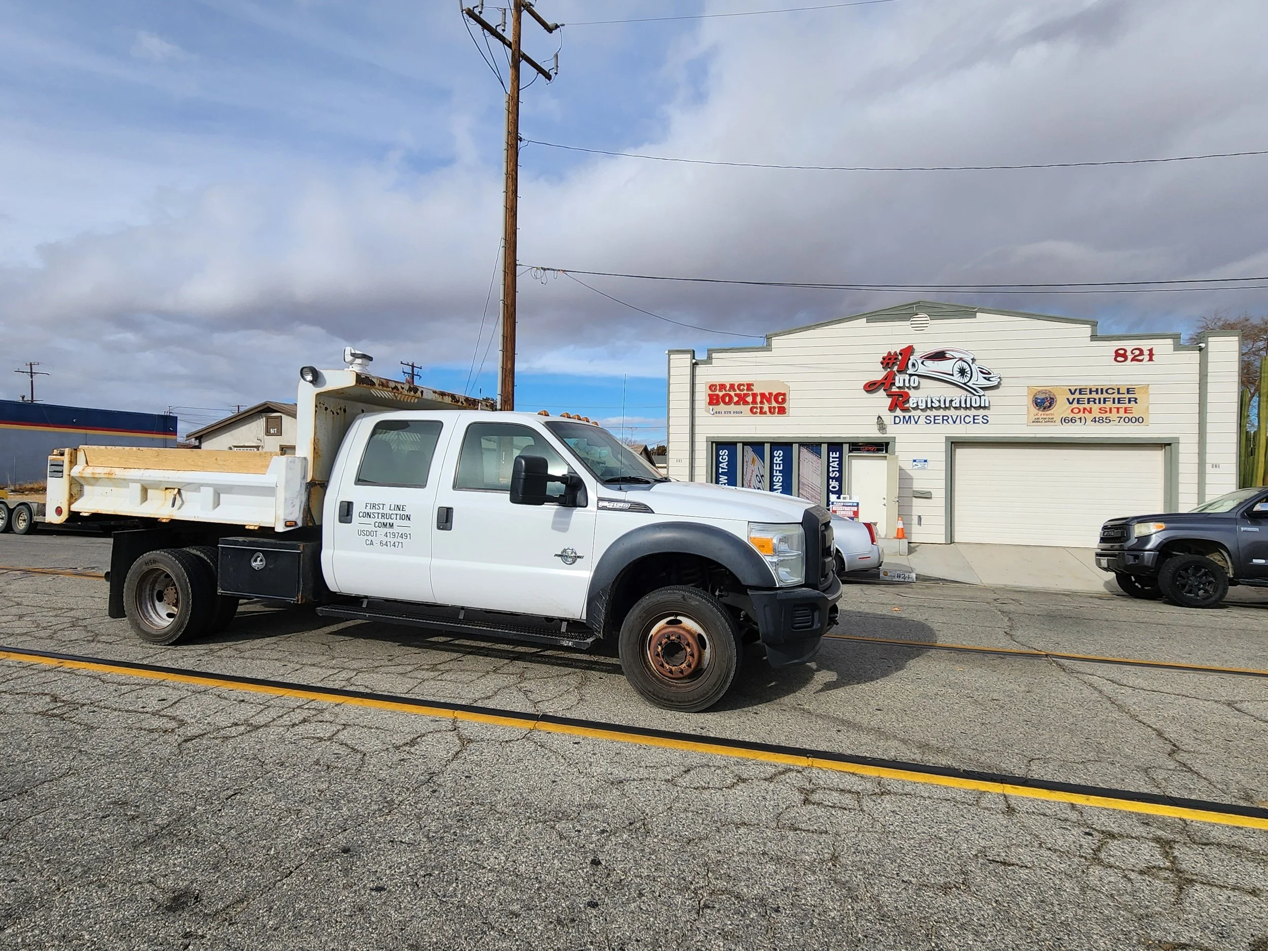 White construction truck parked in front of an auto registration and DMV services shop, with cars parked beside it and utility poles in the background.