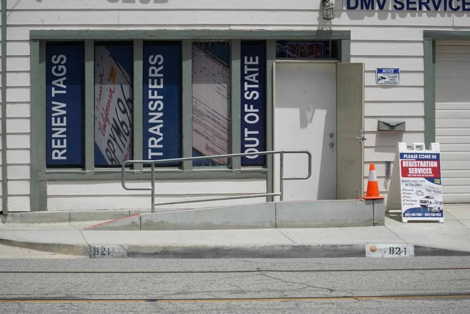 Exterior of a DMV service building with blue signs indicating services such as renewal, transfers, and out of state, a small sidewalk with railing, a traffic cone, and an A-frame sign for registration services. The building has white siding and a small window with an open sign