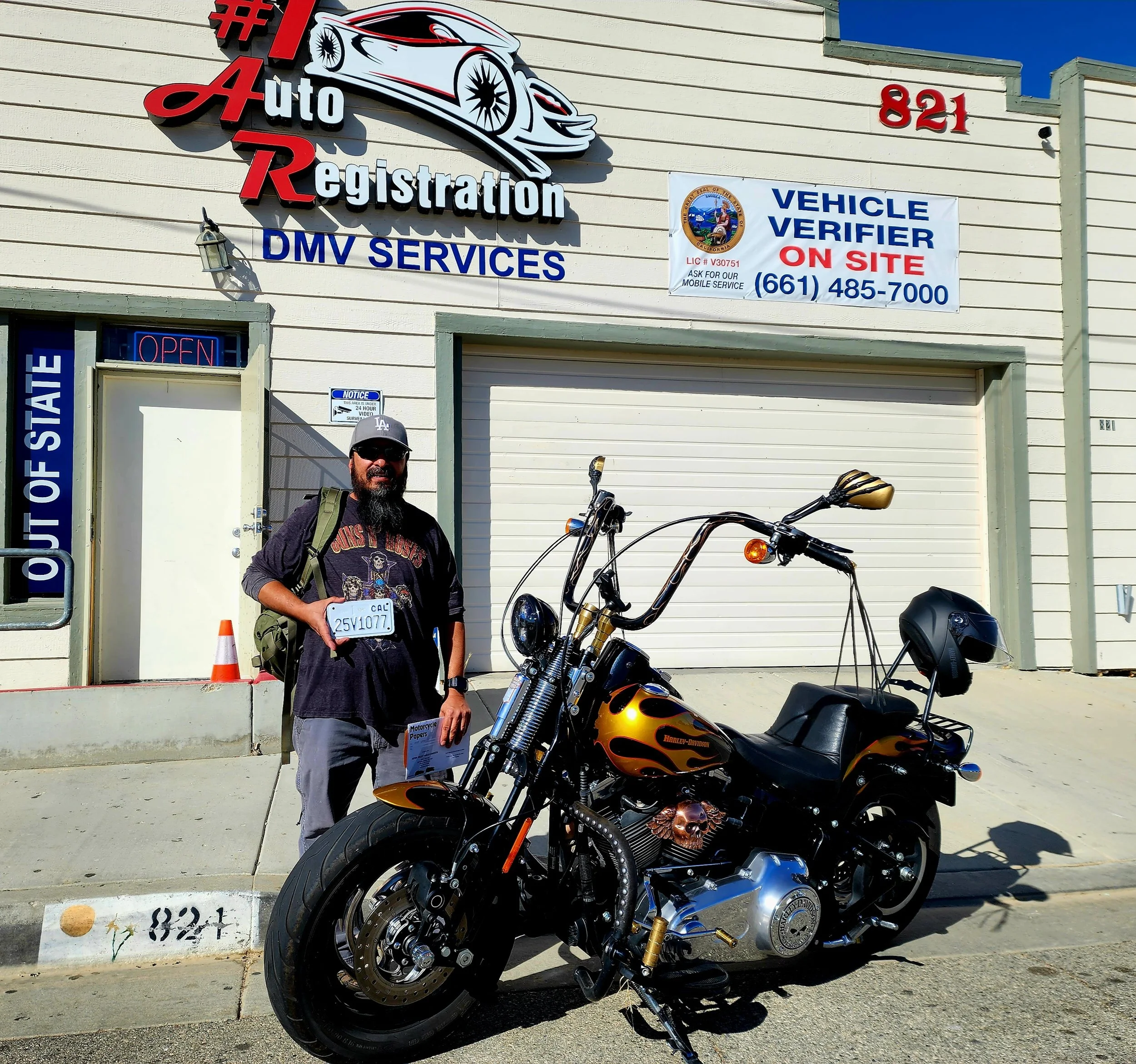 A man with a beard and sunglasses standing next to a black Harley-Davidson motorcycle outside a DMV service building with signs for auto registration and vehicle verification.
