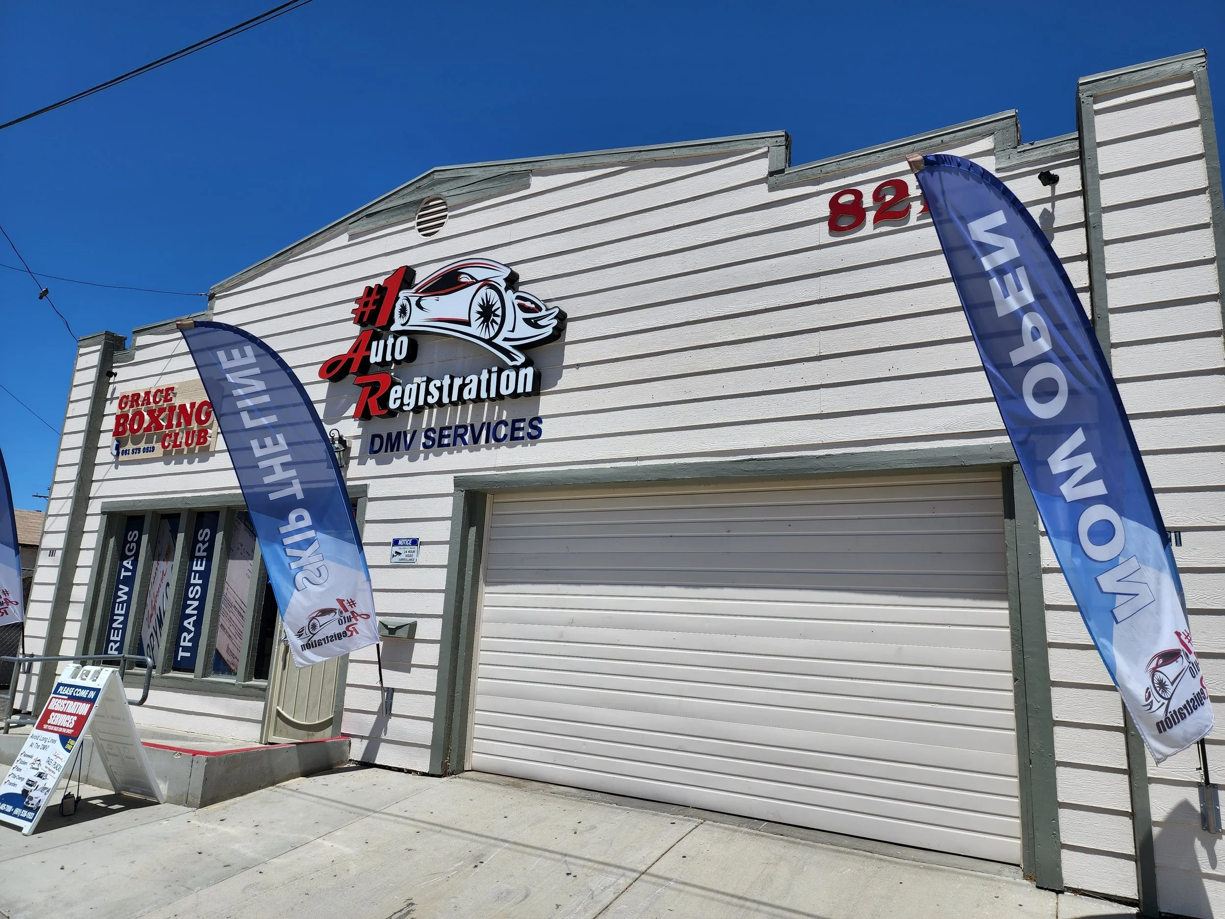 Auto registration shop with flags advertising DMV services, a sign for Grace Boxing Club, and a garage door on a white building with the number 82.