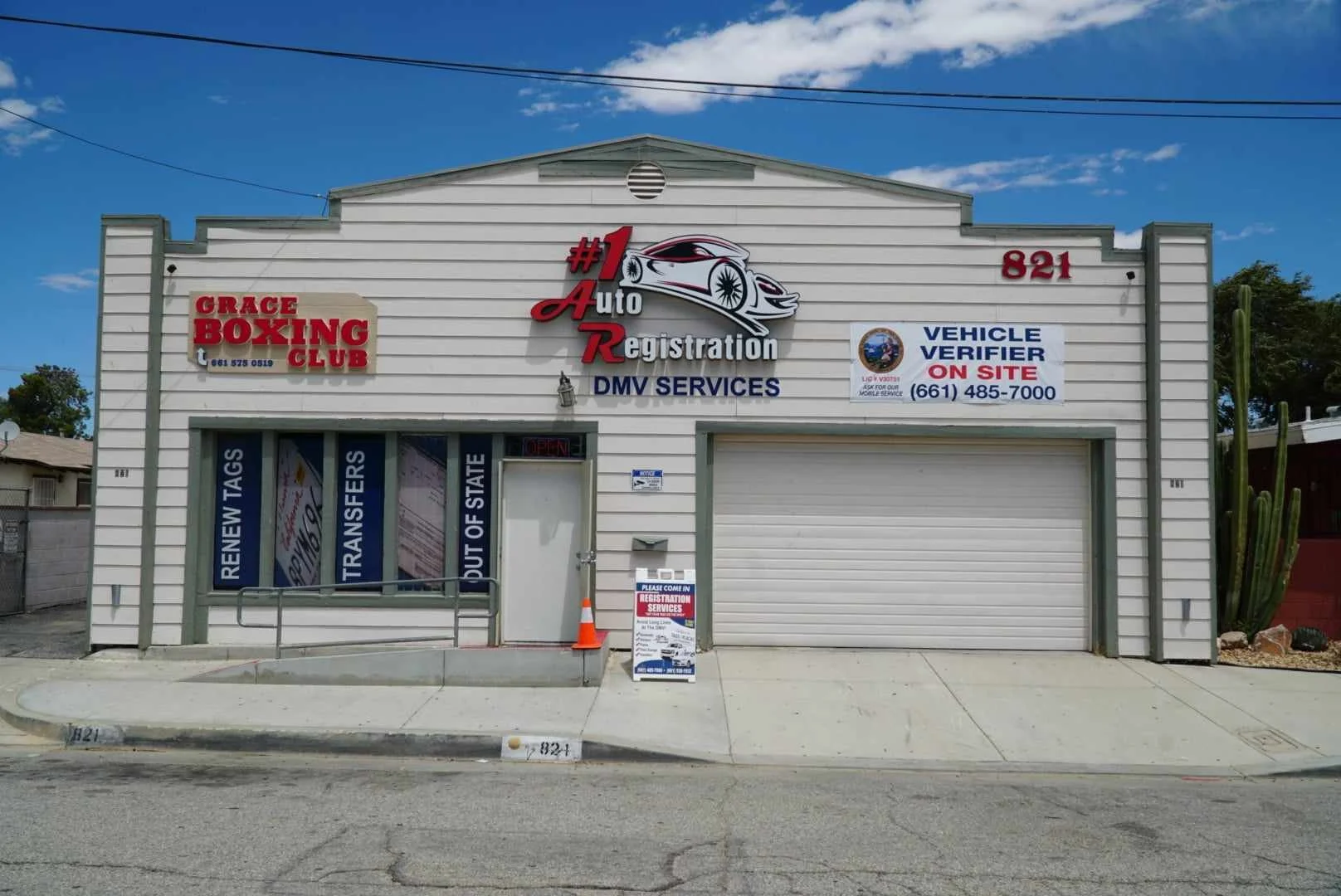 Auto registration and DMV services building with signs for vehicle verification, license renewals, and transfers. The building is white with gray trim, has a roll-up garage door, a pedestrian entrance with an 'Open' sign, a cactus plant on the right, and a small sign in front near the sidewalk.