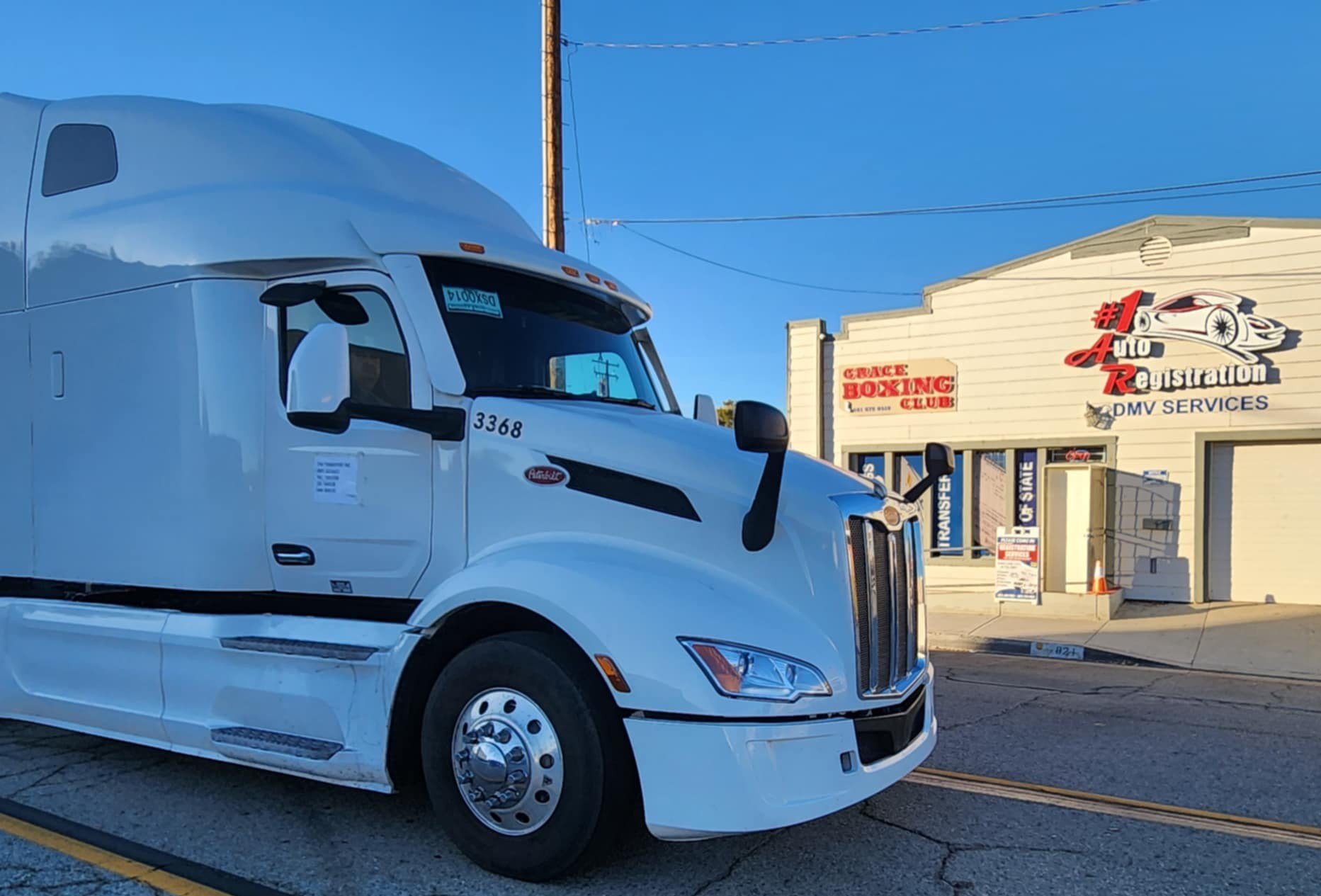 White semi-truck parked outside a building with signs, including '#1 Auto Registration' and 'Grace Boxing Club.'