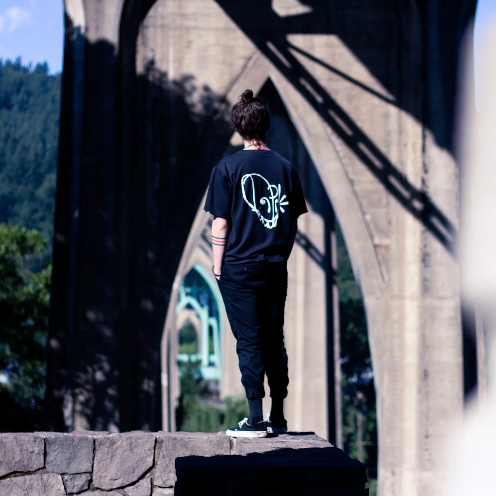 A person with dark hair in a bun, wearing a black t-shirt with a graphic face on the back, black pants, and sneakers, standing on a ledge beneath a stone bridge with shadows cast by the bridge structure.