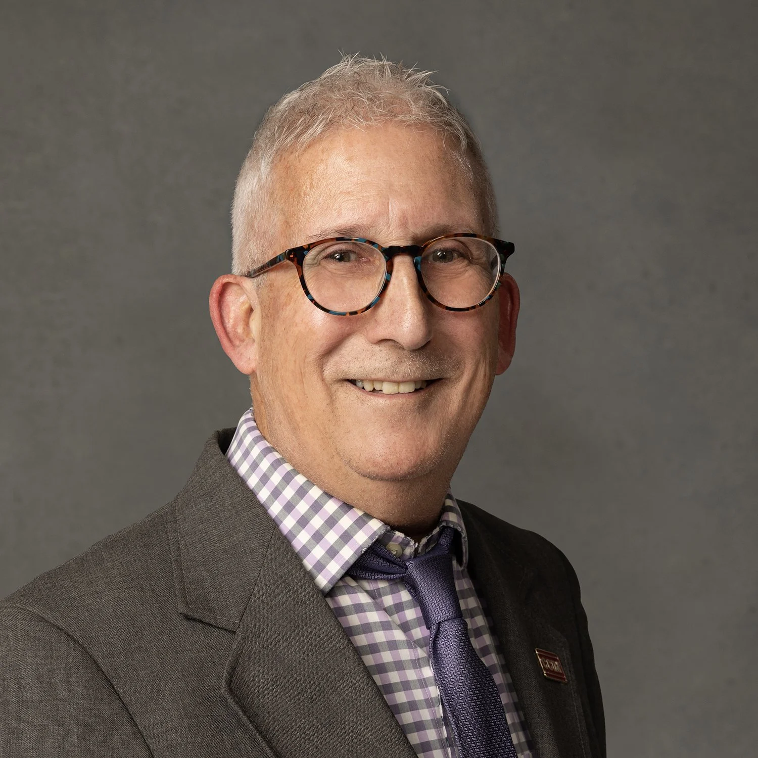 Smiling man wearing glasses, a gray suit, checkered shirt, and a tie, against a gray background.