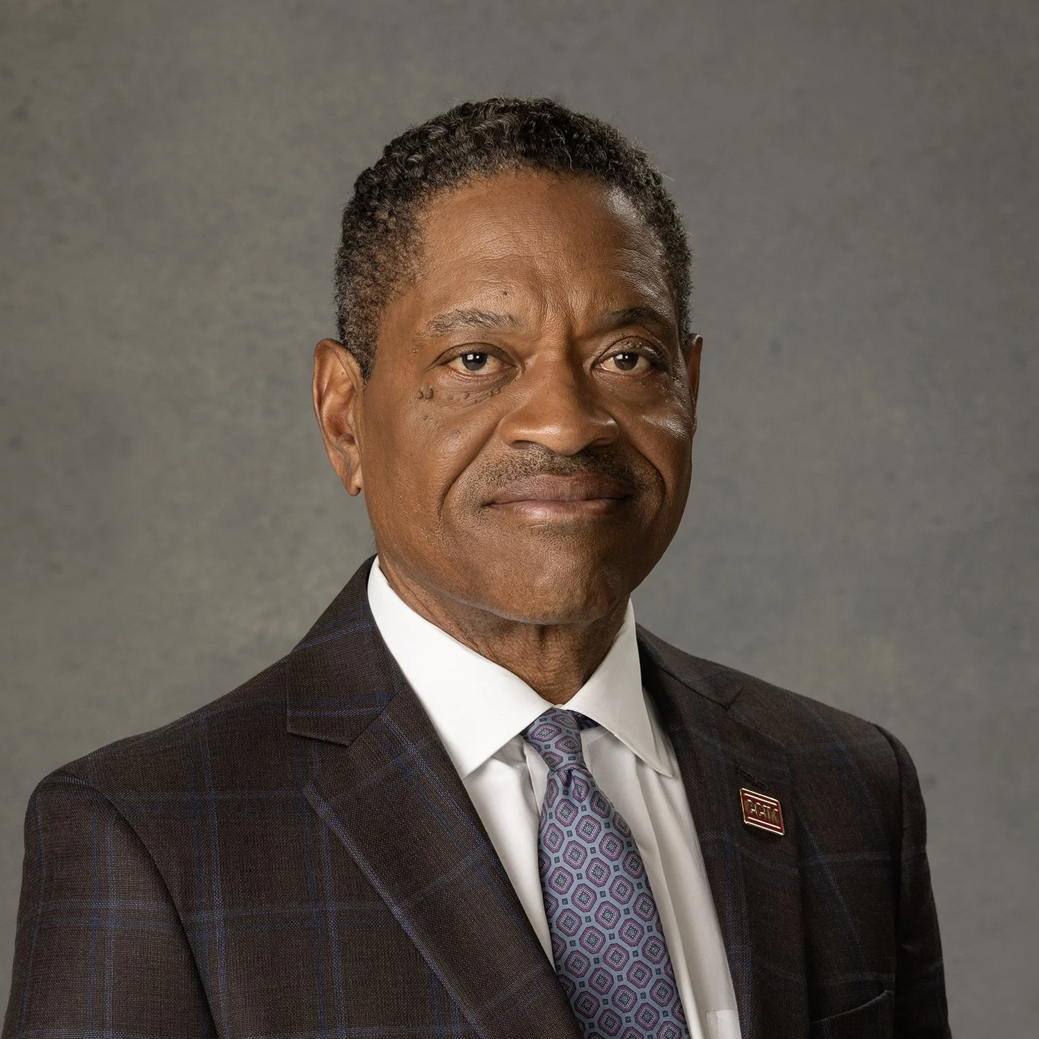 A man in a suit and tie with a neutral expression, against a plain background.