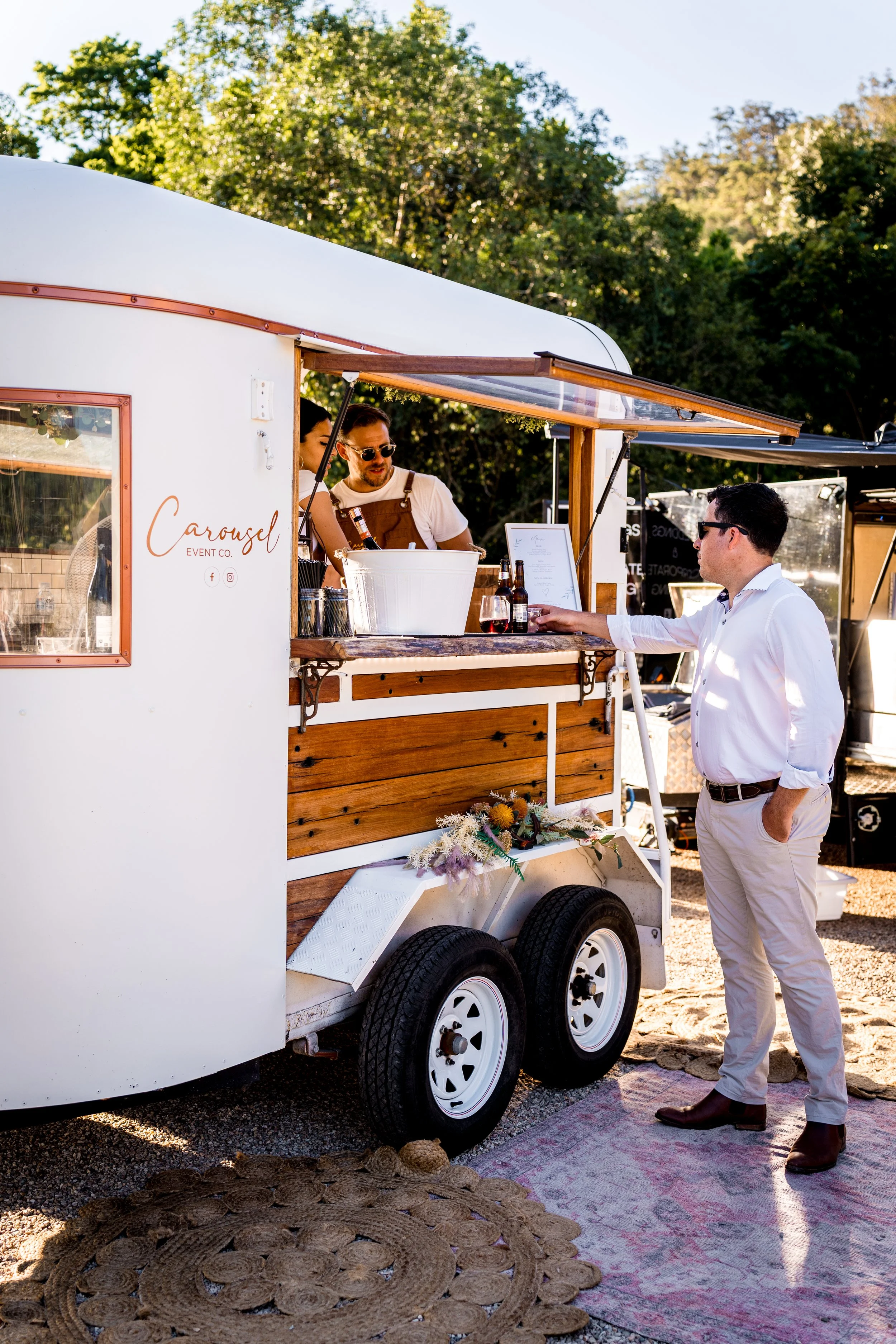 A man in white shirt and beige pants ordering drinks from a small white and wooden food truck named 'Carousel Event Co.' with two people inside. The truck has flowers attached below the window and is parked outdoors with green trees in the background.
