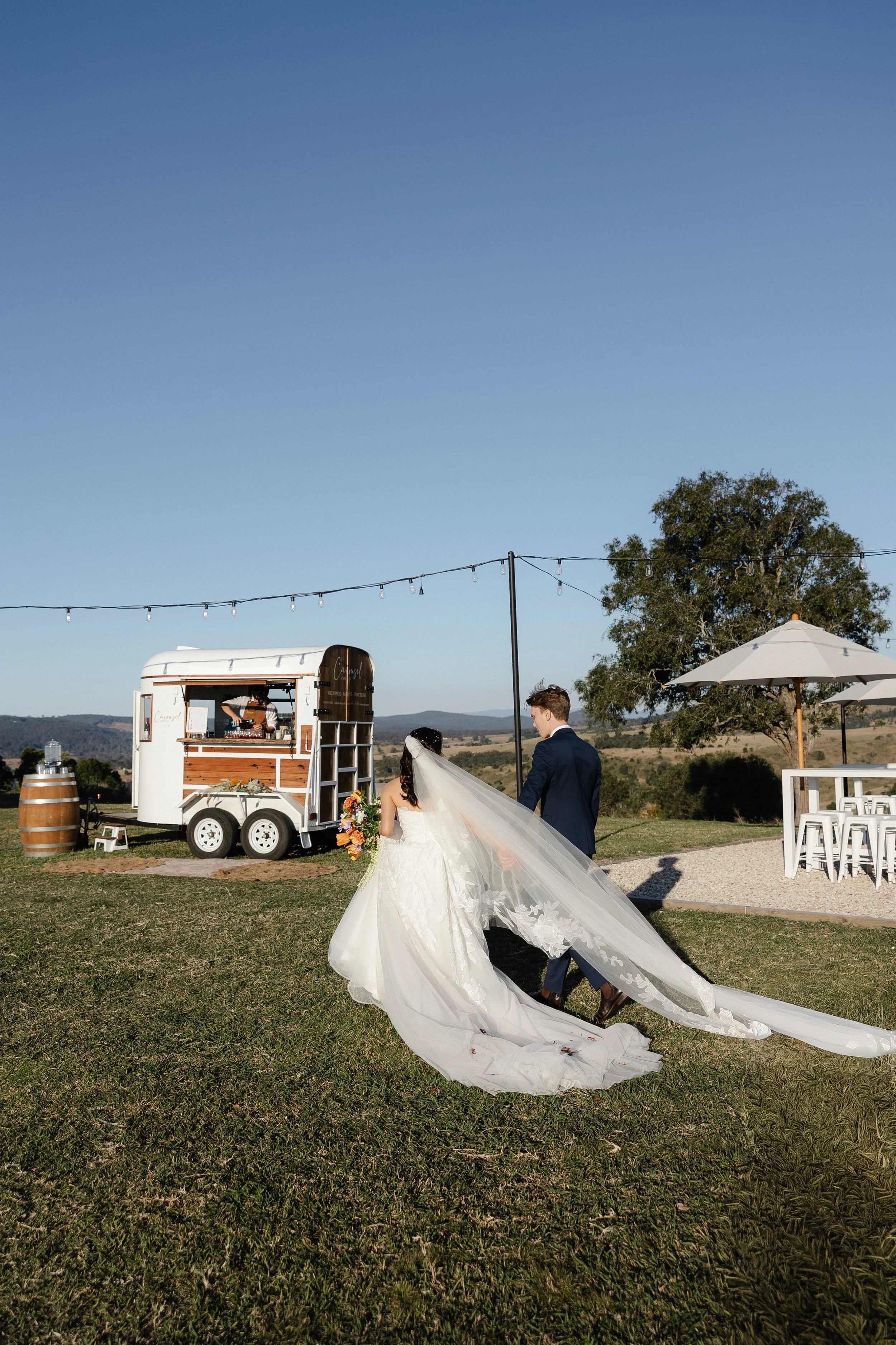 A bride and groom walking together on a grassy field at an outdoor wedding reception in front of a mobile horse float bar.