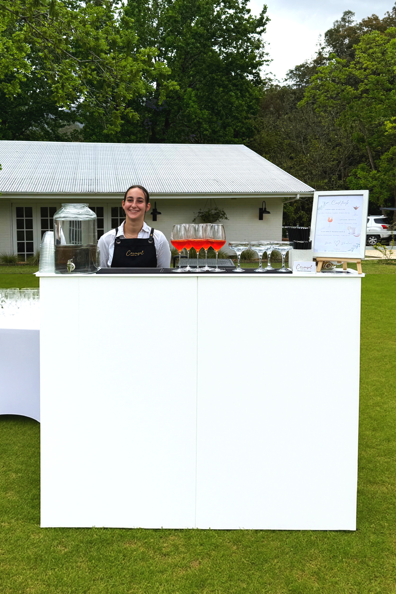 A smiling woman standing behind a white outdoor bar setup with four glasses of red cocktail, a water dispenser, and a framed menu, outdoors on a grassy area with trees and a house in the background.