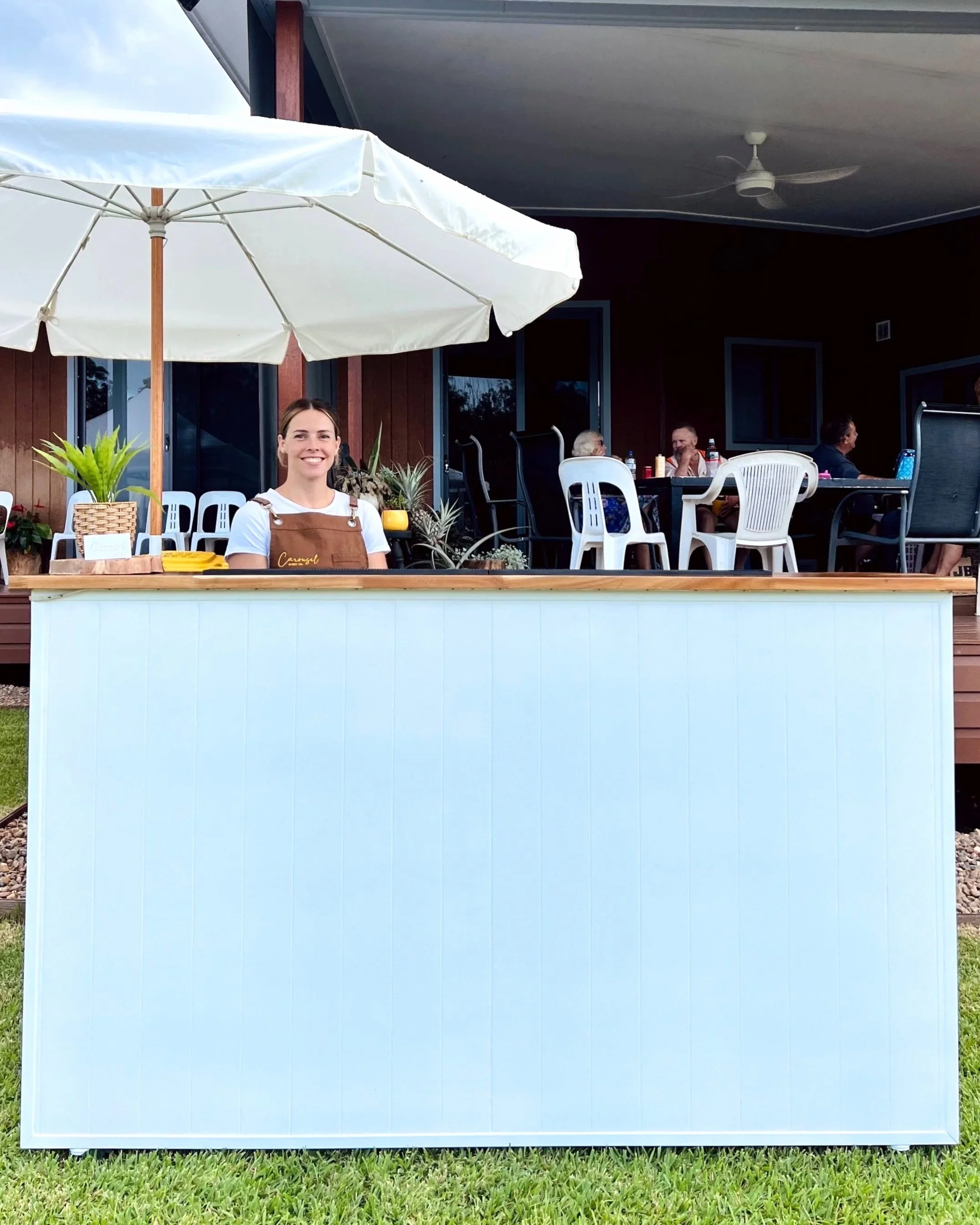 A woman in an apron standing behind a white mobile bar, with a large white patio umbrella overhead and a patio area with chairs and people at an event