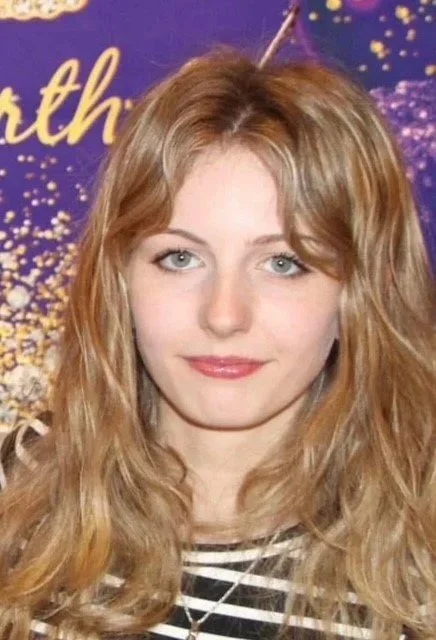 A young woman with long wavy blonde hair and blue eyes, smiling slightly, wearing a black and white striped shirt and a necklace, standing in front of a purple backdrop with gold confetti and partial gold lettering.