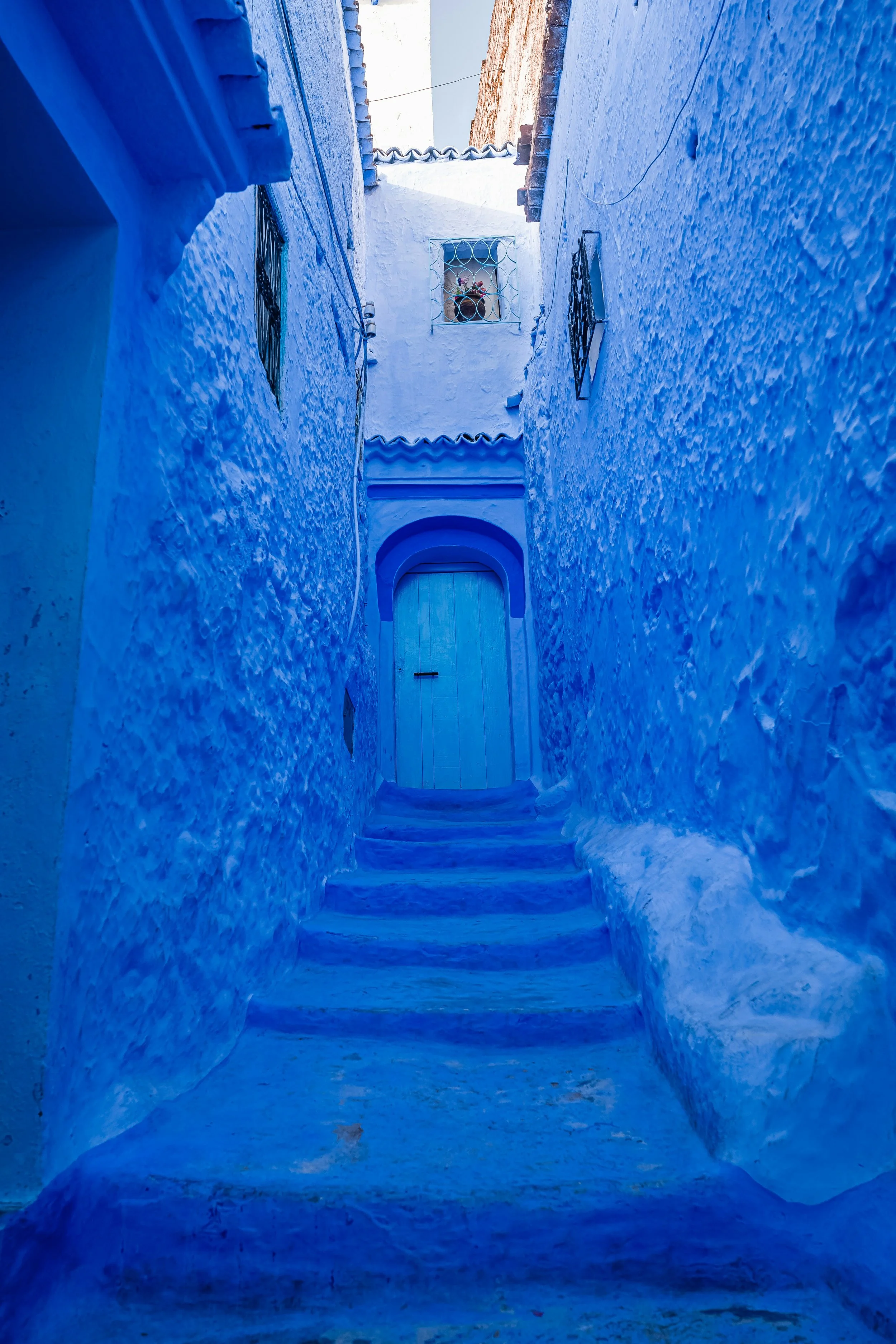 Narrow alleyway with textured white and blue painted walls, a small door at the top of steps, and a flower window on the second floor.