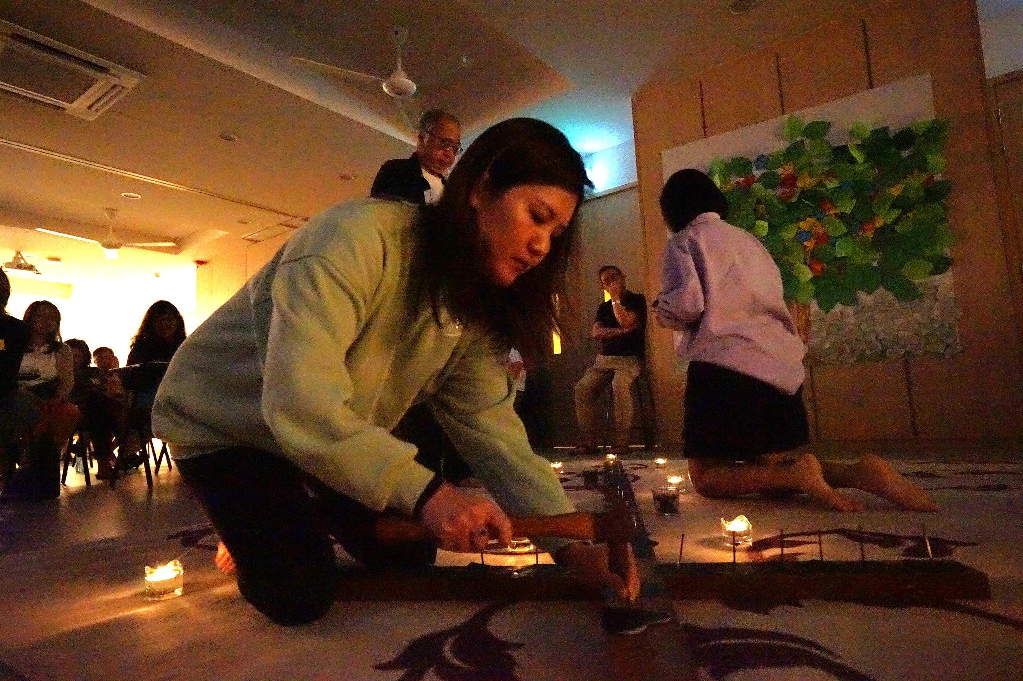 People kneeling on the floor surrounding candles and a large painting of a tree, participating in a ceremony or meditation in a dimly lit room.