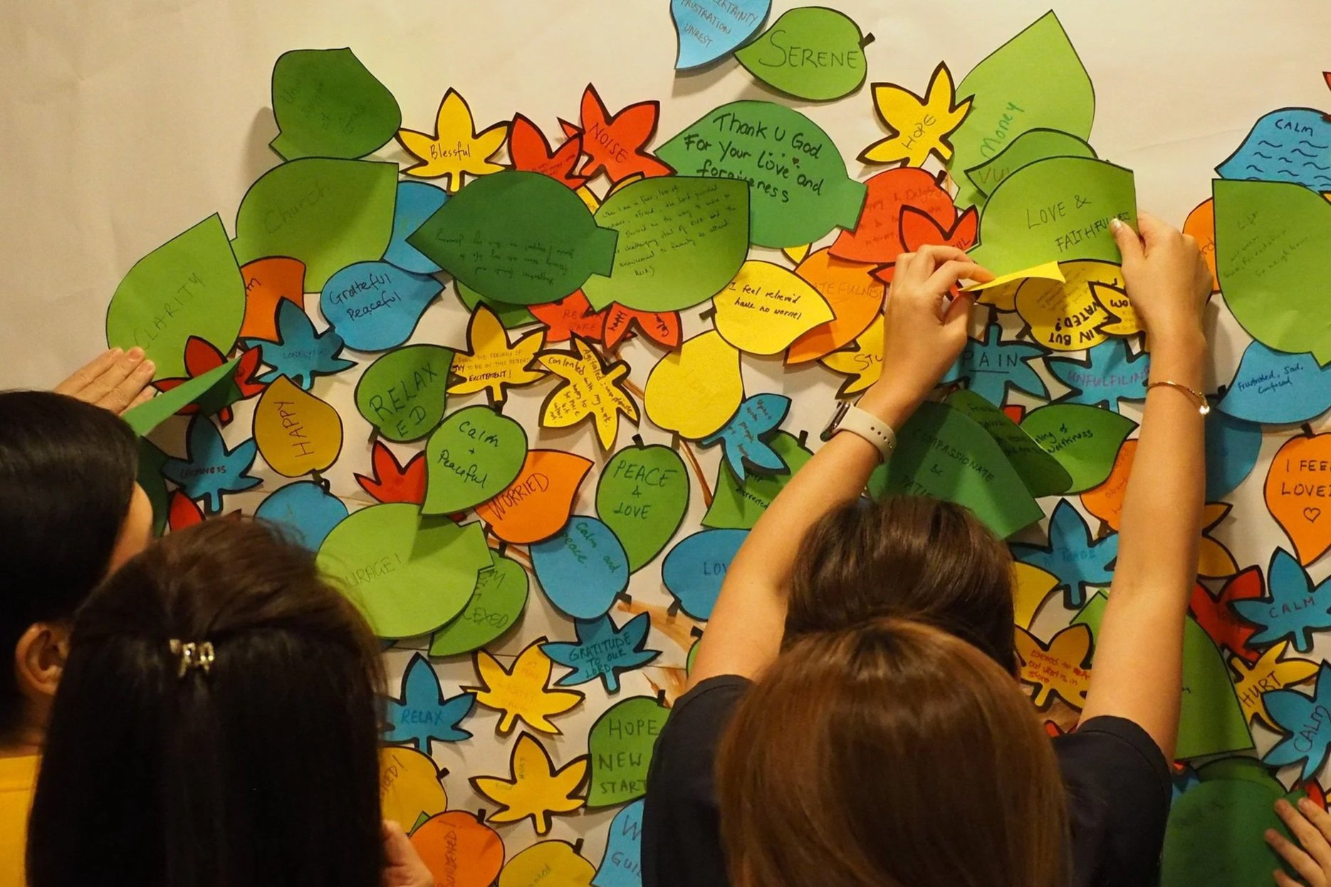 People placing colorful paper leaves with wishes and messages on a wall for a communal display.