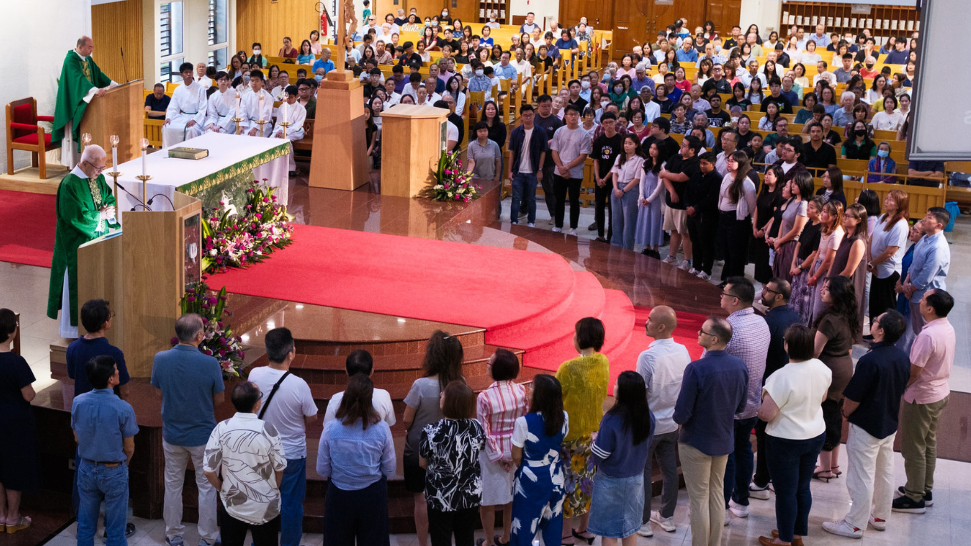 A large congregation gathered inside a church during a service. Two priests are at the altar, one reading from a lectern, with a cross and floral arrangements nearby. The congregation includes men, women, and children, some wearing masks, standing and attentively participating.
