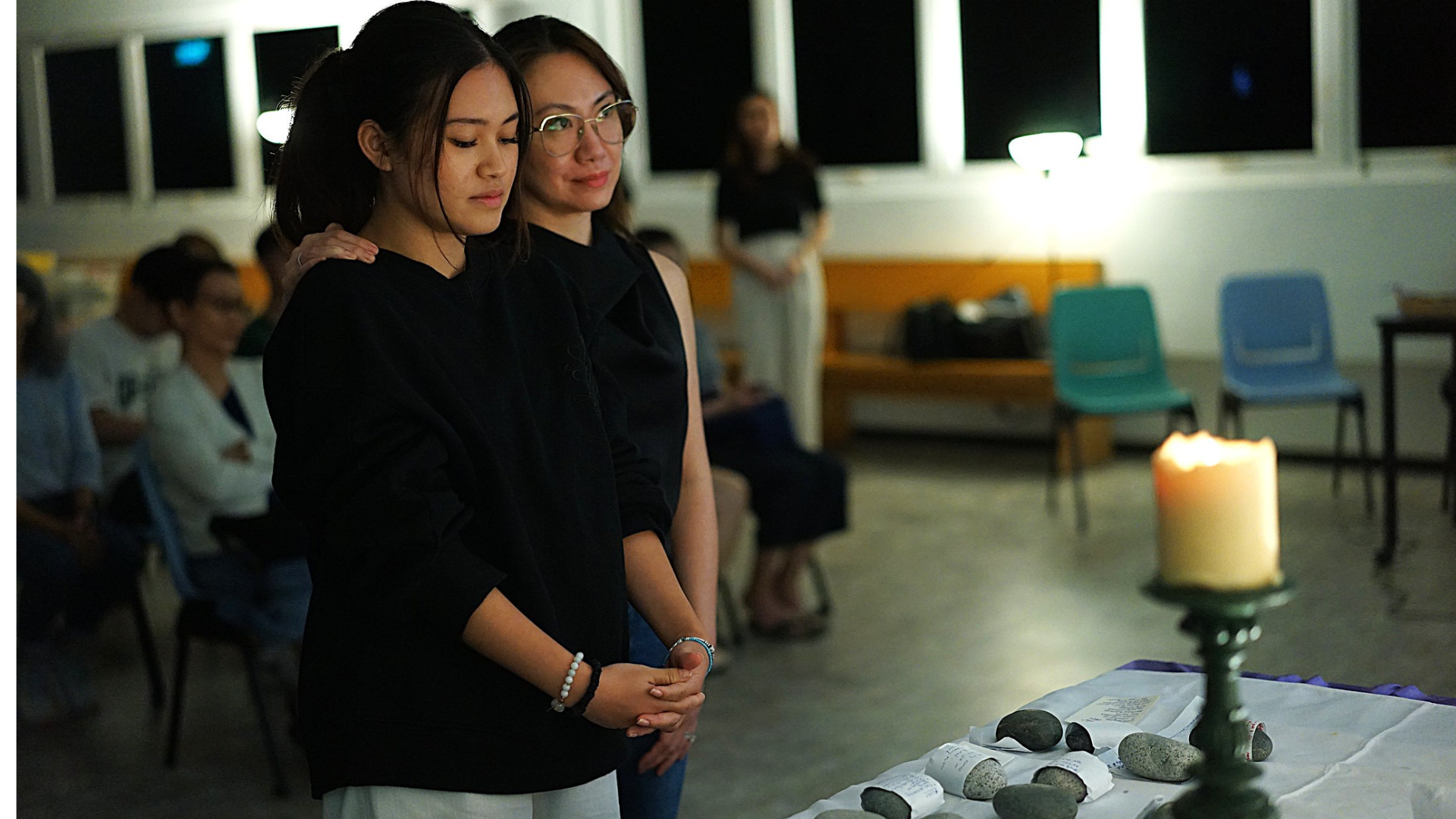 Two young women standing in prayer or reflection during a candlelit ceremony, with rocks and papers on a table in front of them, in a dimly lit room with seated people in the background.