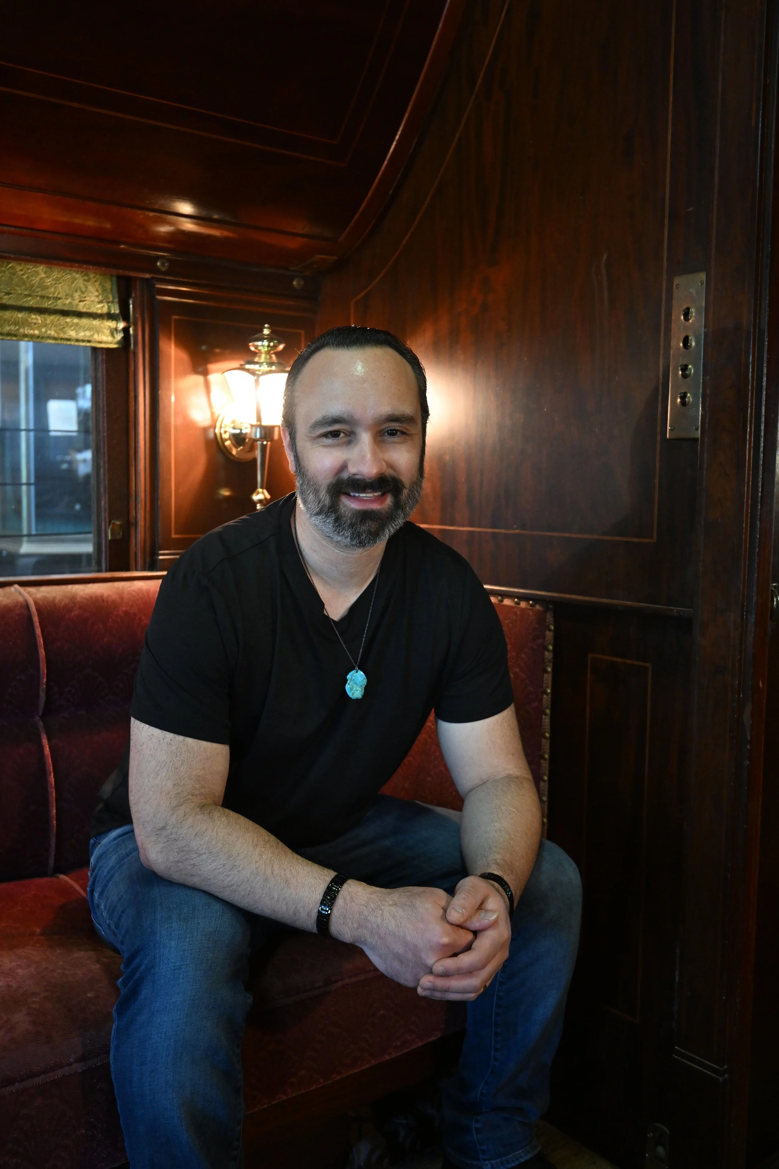 A man with a beard and mustache sitting on a red velvet bench in a warmly lit room with wooden walls, wearing a black shirt and blue jeans, smiling at the camera.