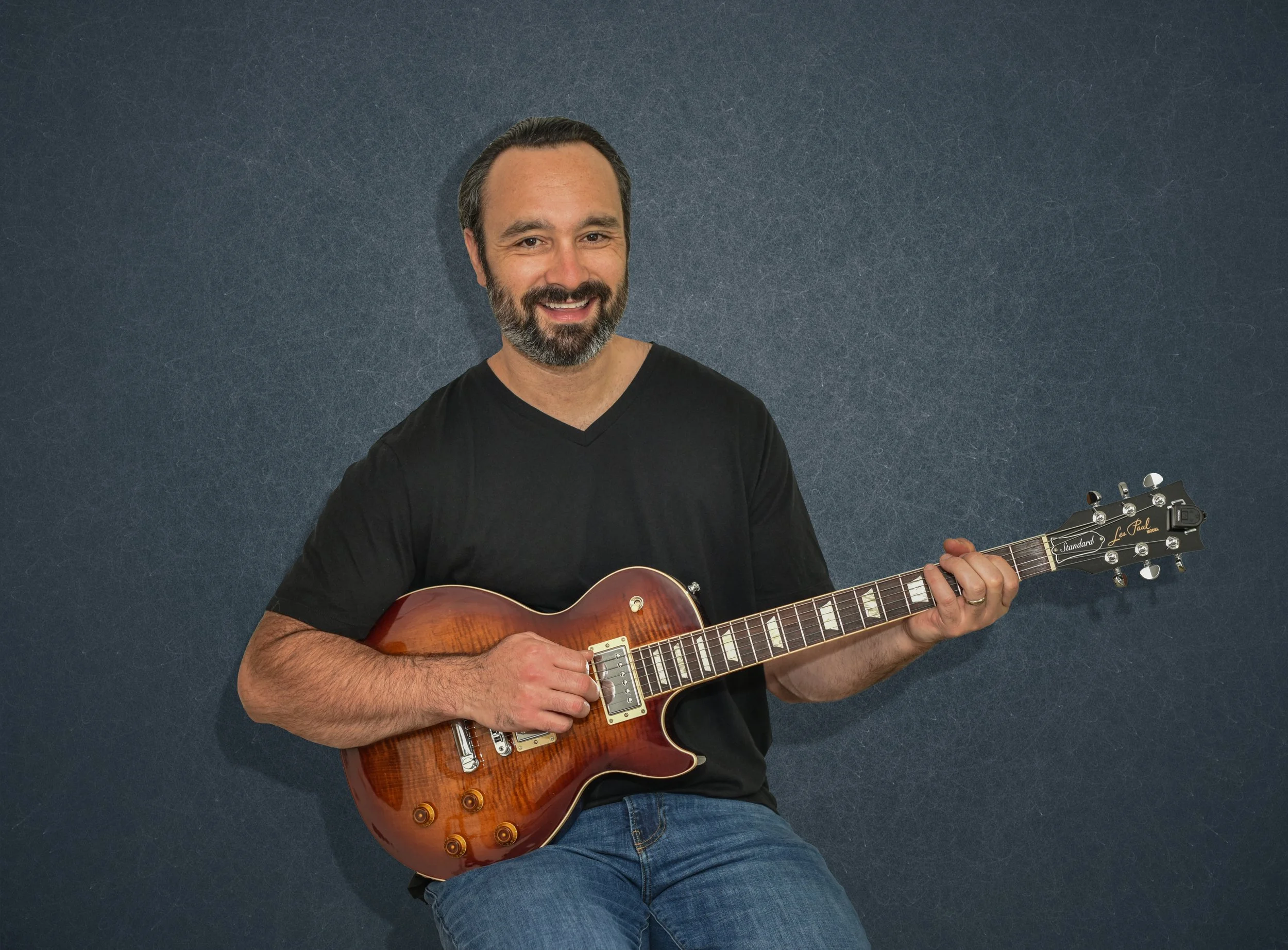 A man with dark hair and a beard smiling while holding an orange sunburst electric guitar in front of a dark, textured background.