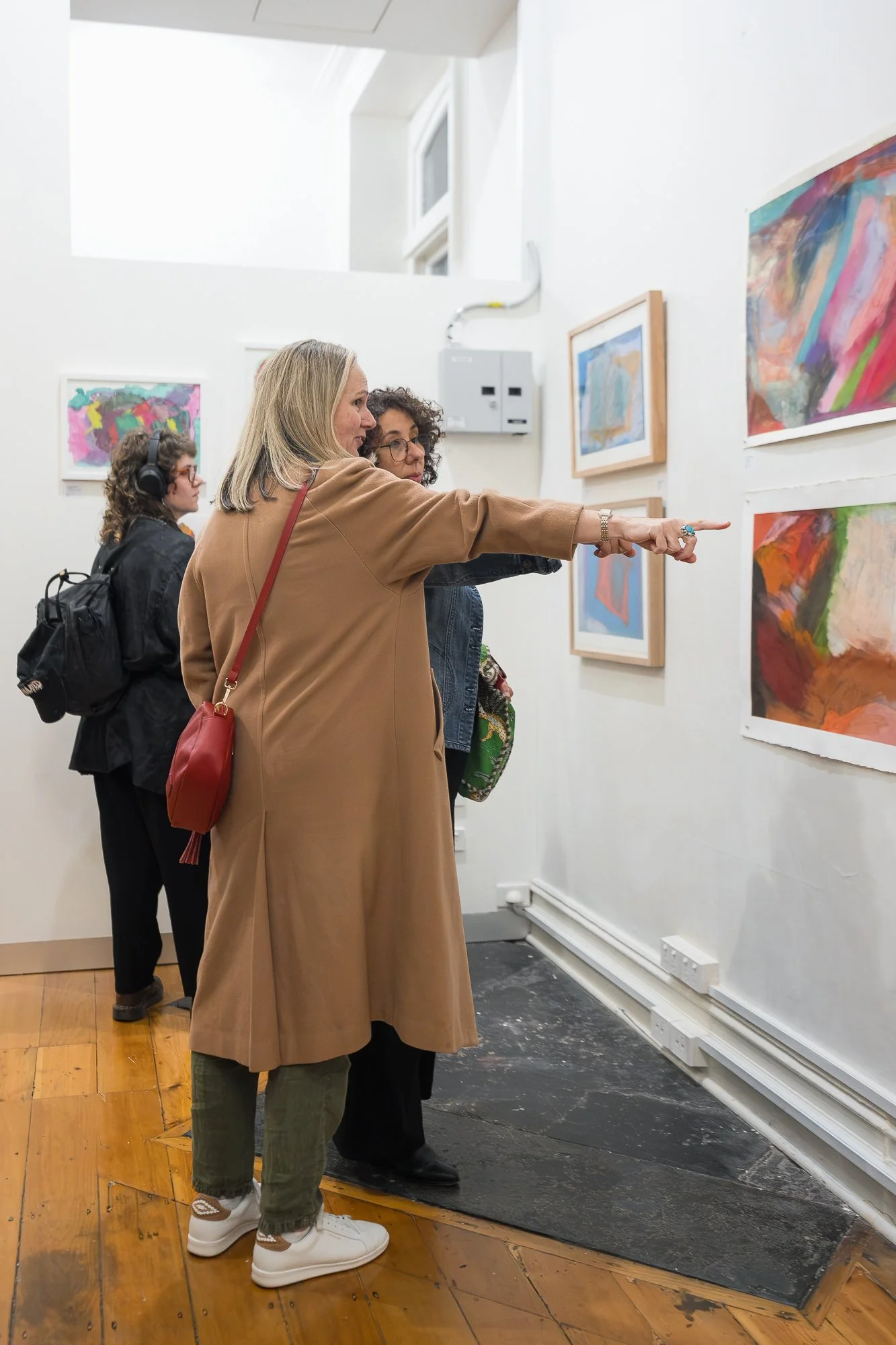 A woman in a camel coat points out a specific work to her companion among a wall of bright, colourful paintings.
