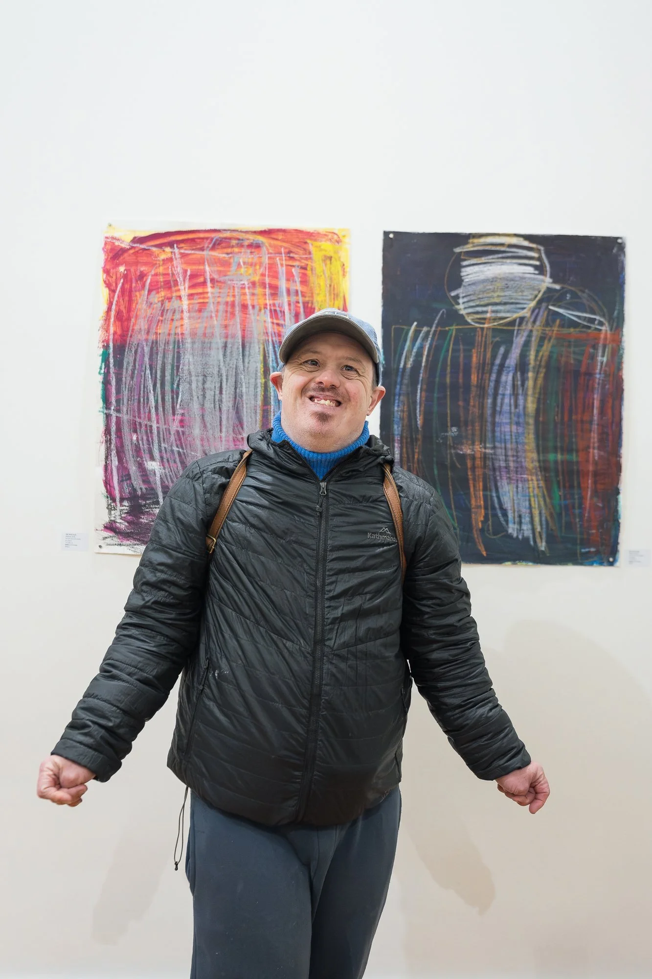 A man in a cap grins broadly, arms out, standing in front of the same two large expressive works — pure joy.