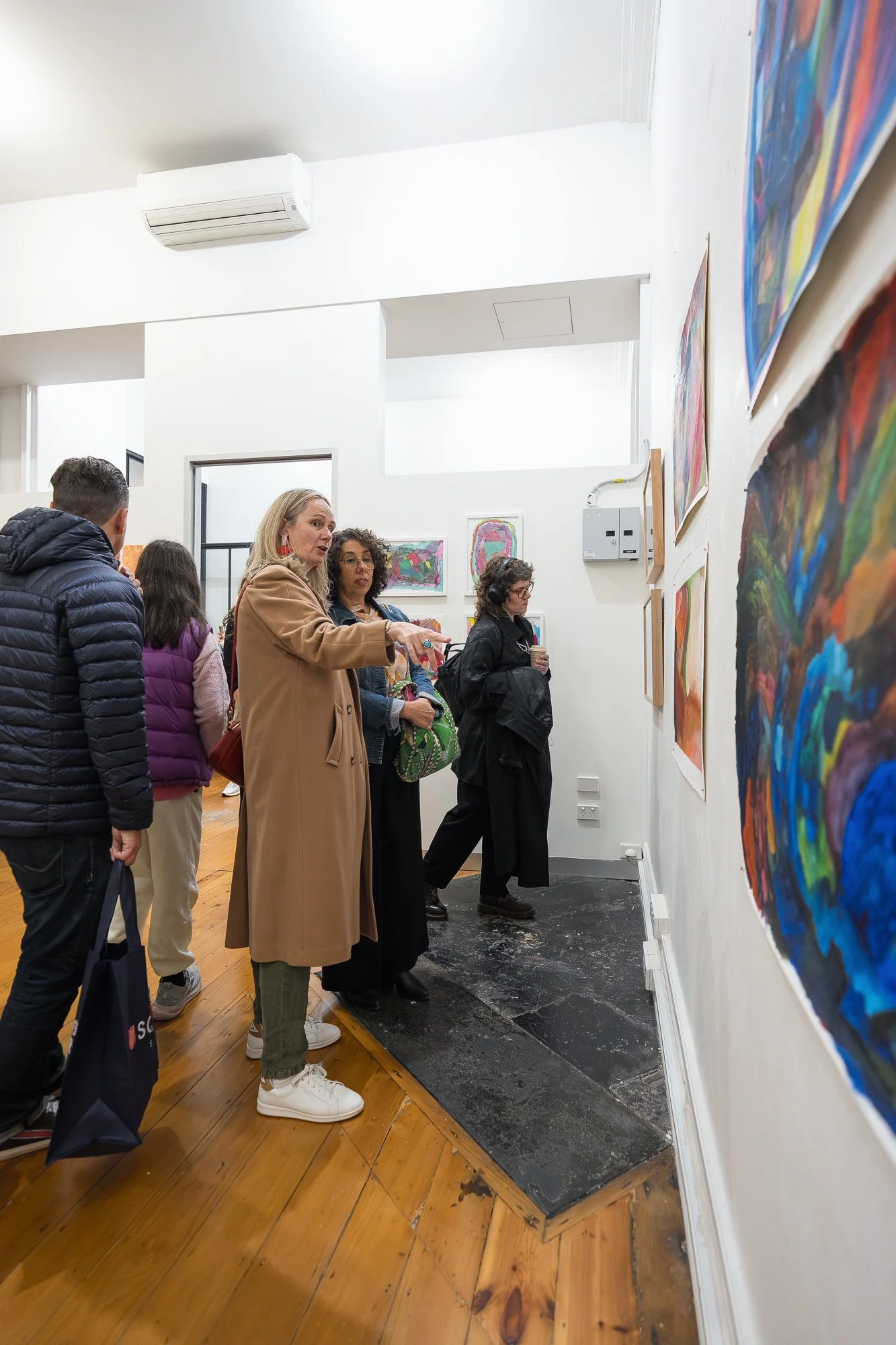A woman in a camel coat gestures toward the works as she guides another visitor along a wall of colourful paintings.