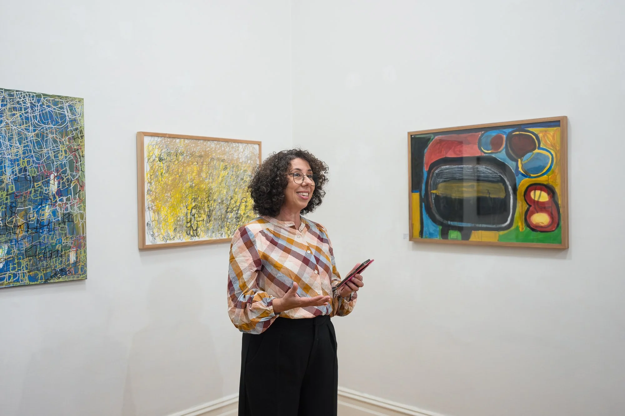 A woman in a colourful check blouse gestures expressively as she speaks, surrounded by the artists' works.