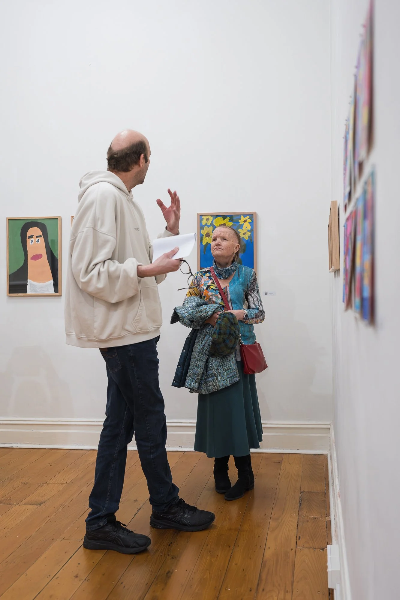 Man with handout sheet talking to a woman in a patterned jacket, standing near a portrait painting and a sunflower still-life on the gallery wall, with rows of small framed works visible to the right.