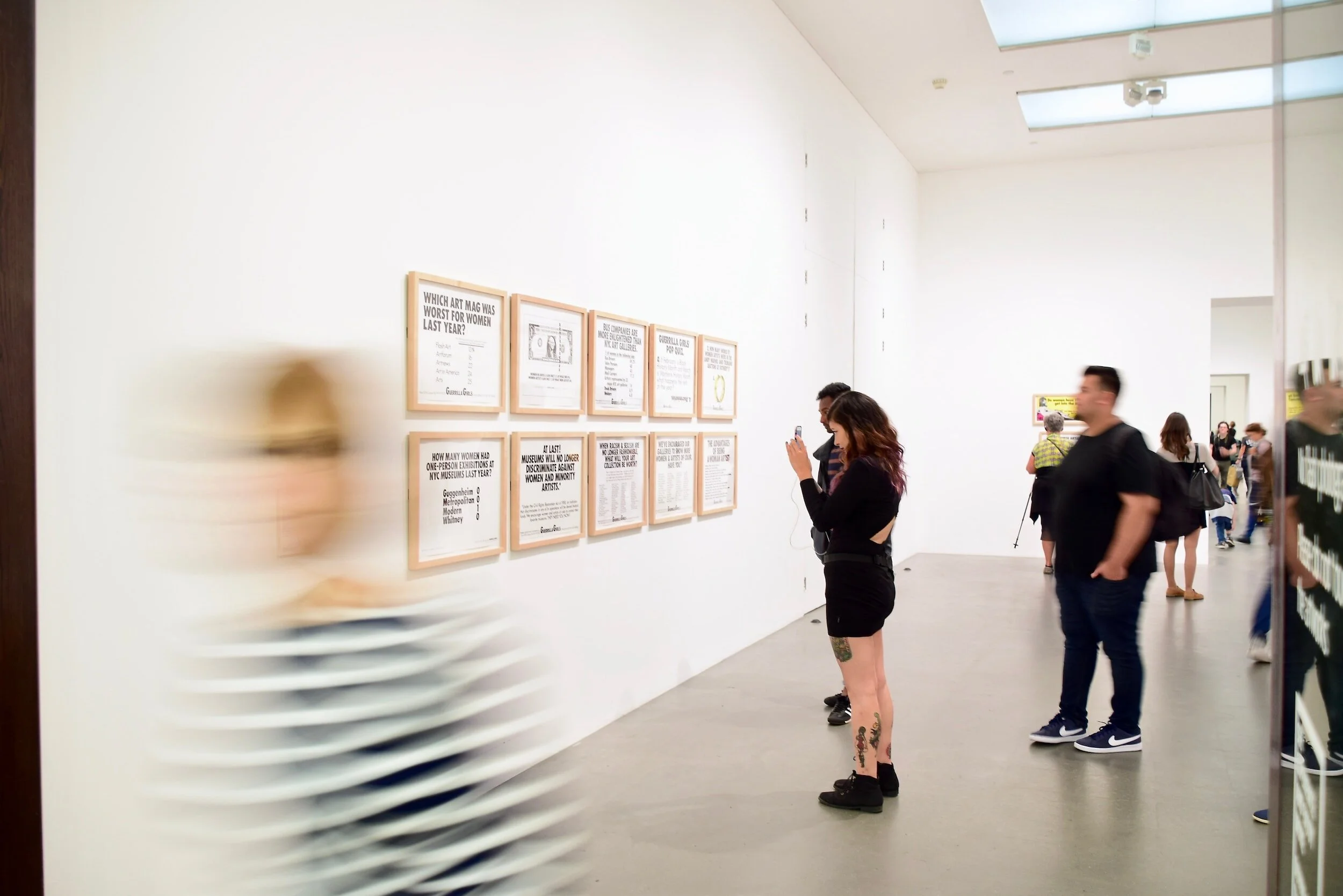 Image of a light-filled gallery space, with four frames artworks and people looking at the art and moving through the space.