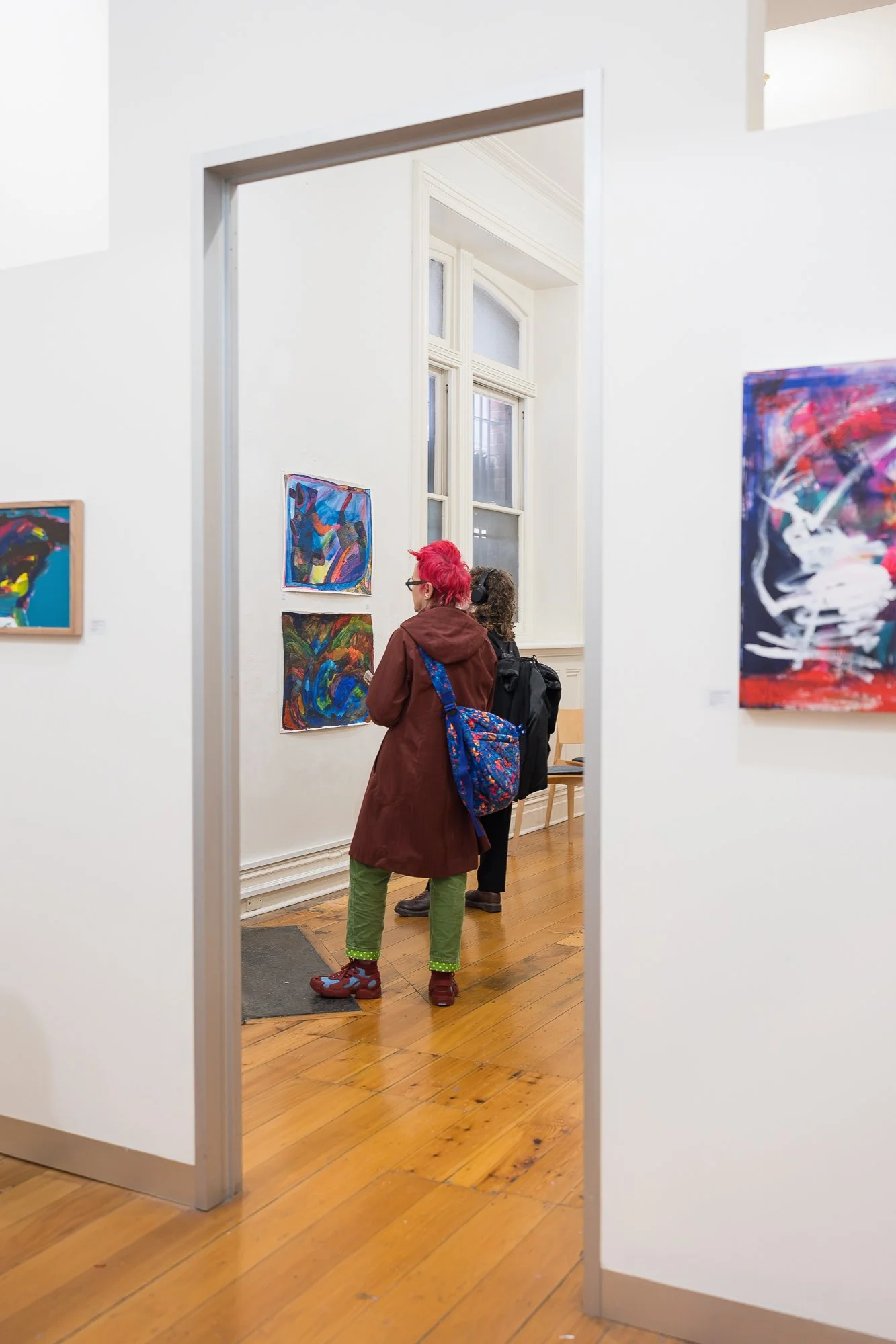 Two visitors are glimpsed through a doorway, quietly looking at small works in an adjoining gallery room.