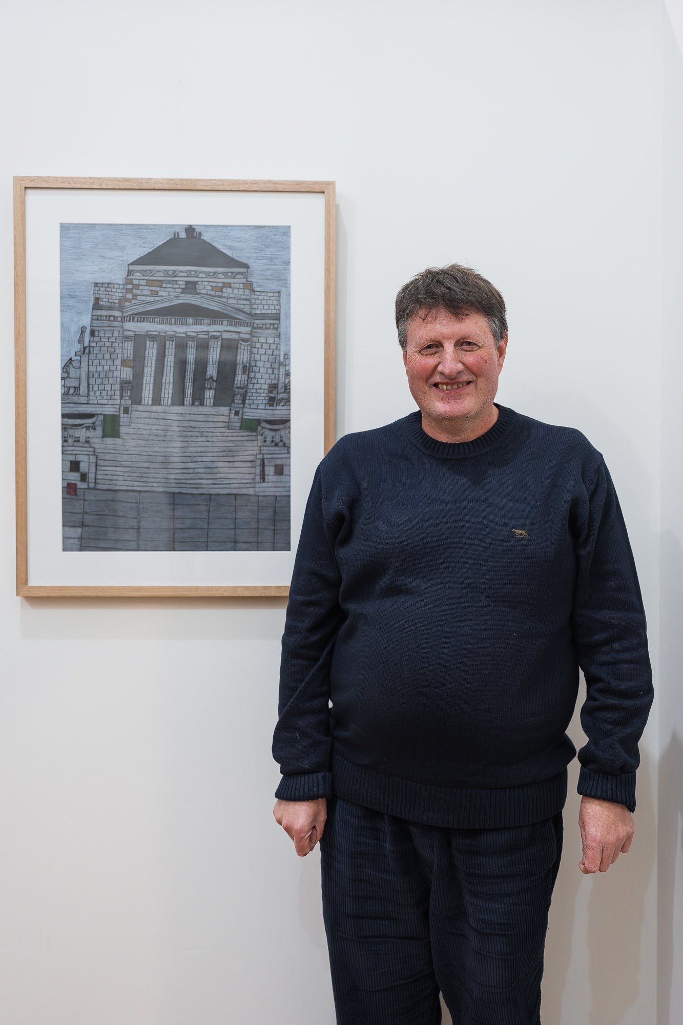 Portrait of a man in a navy jumper standing beside a framed illustration of the Shrine of Remembrance.