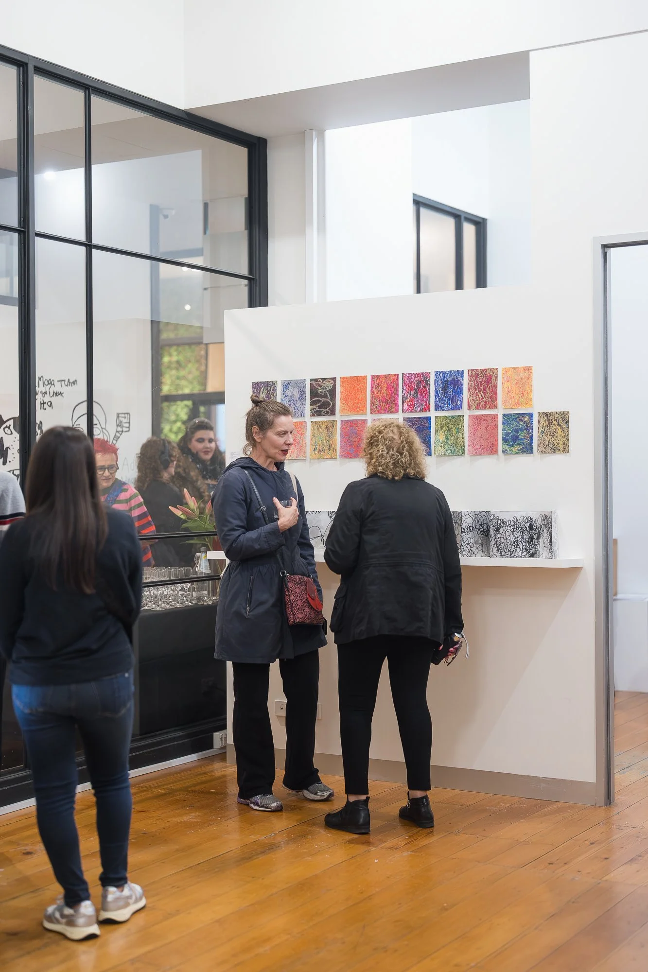 Three people gathered around a grid of small vivid works, deep in conversation in the main gallery space.