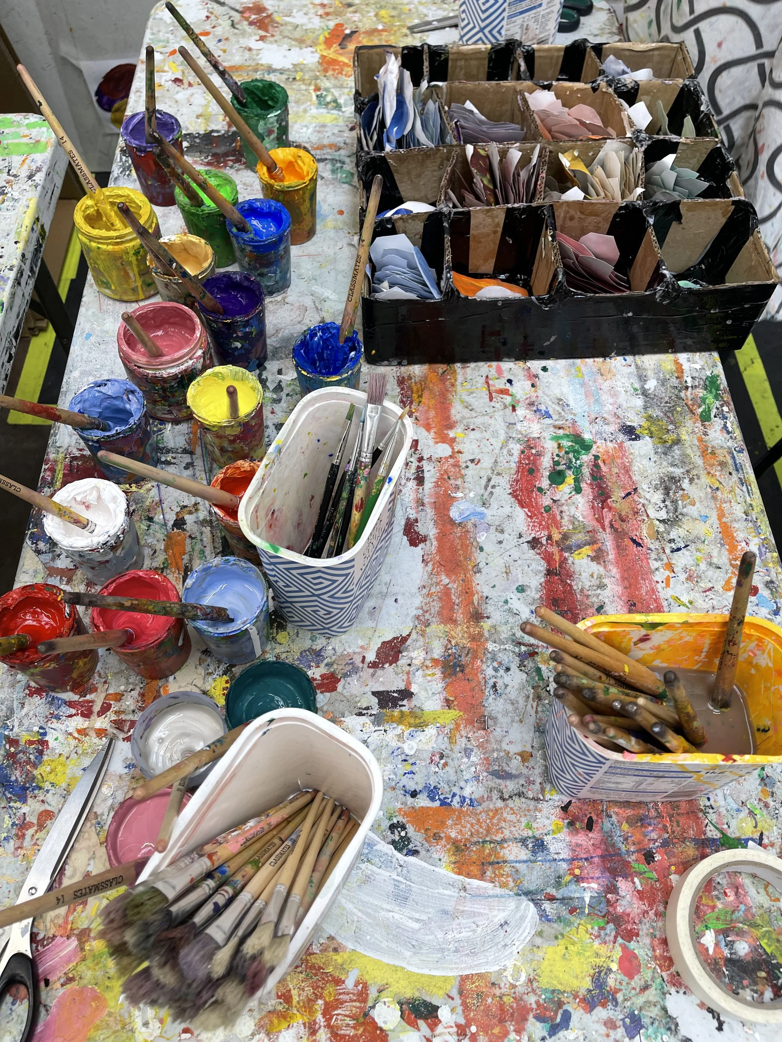 Messy art table with colorful paint jars, brushes, and paper scraps.