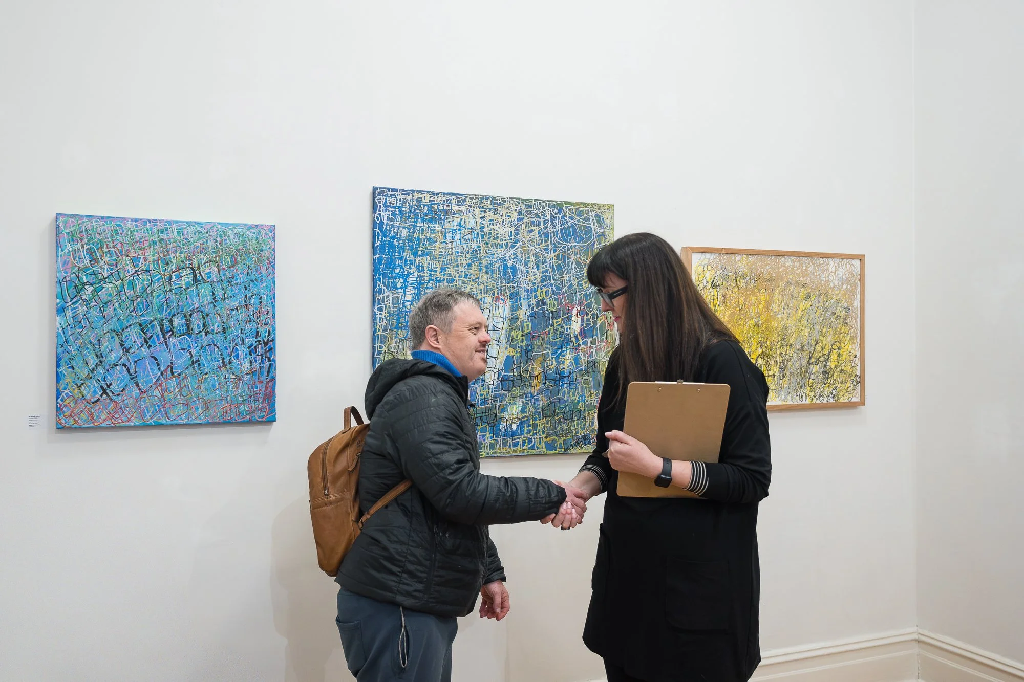 An artist and a gallery representative shake hands warmly in front of three large abstract paintings in blue and yellow.