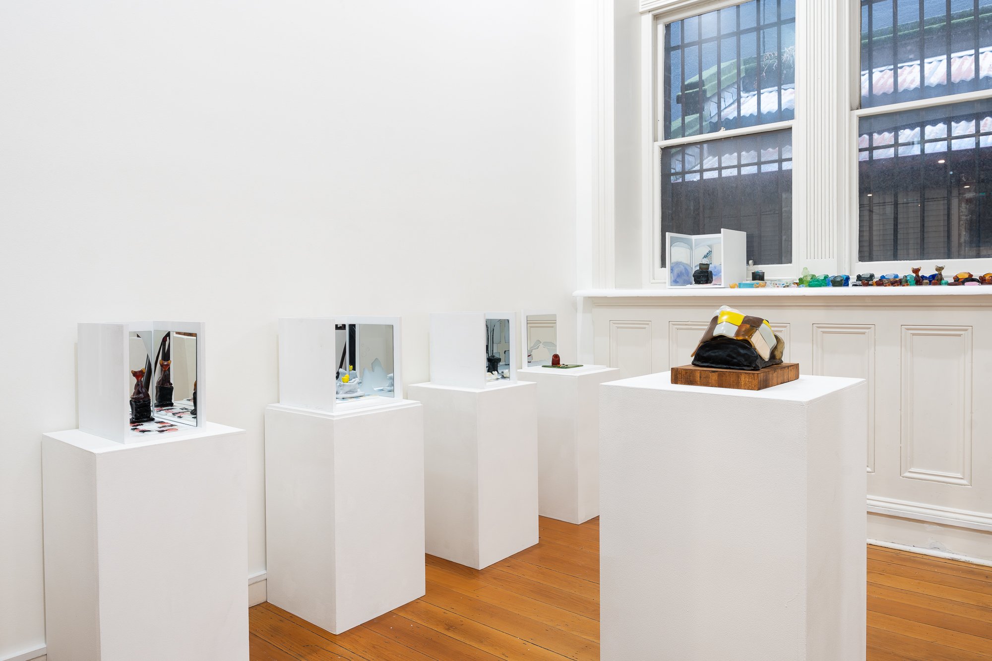 Installation image of glass and wood artworks on white plinths and shelf in a white gallery space with wooden floors
