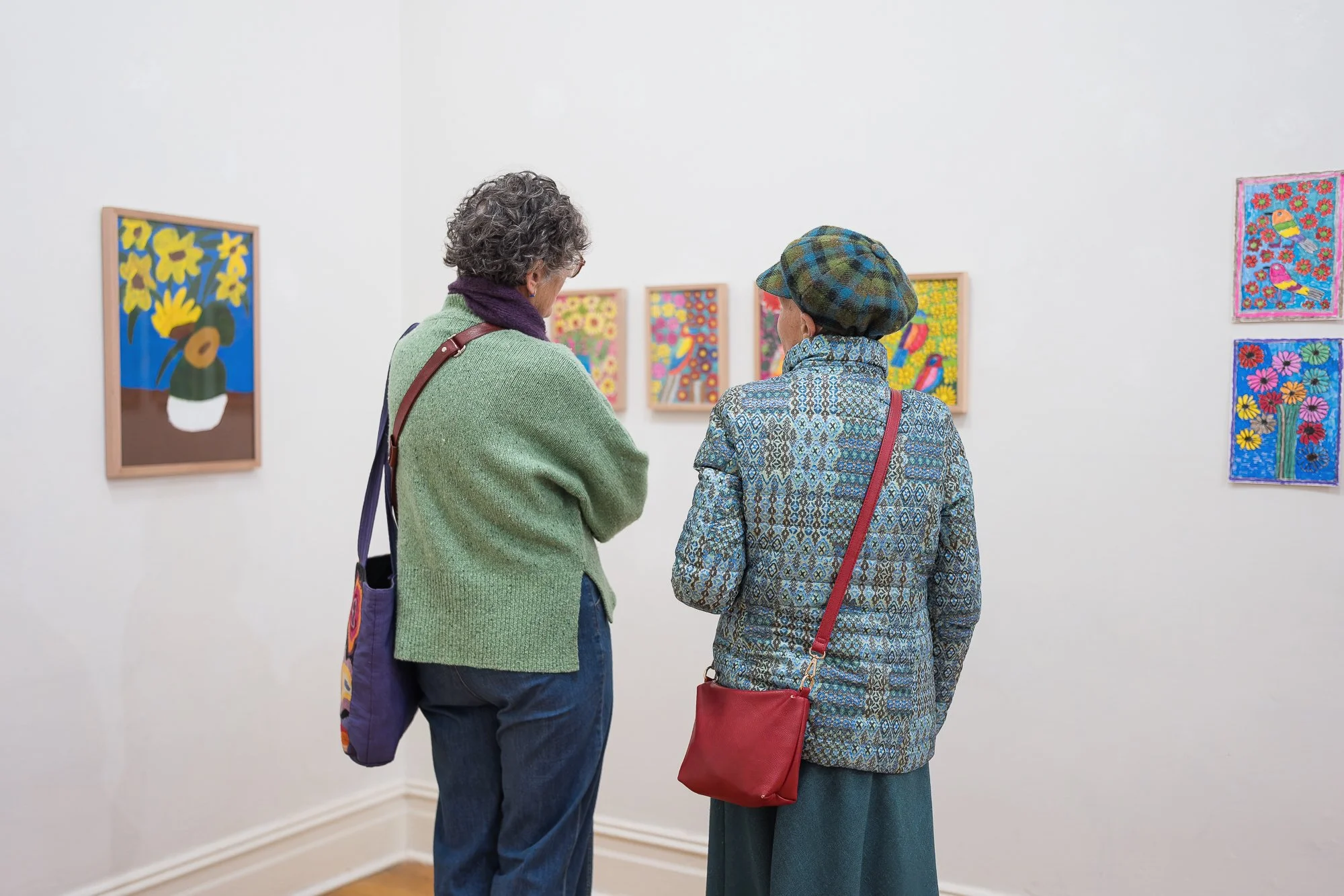 Two visitors viewed from behind, standing in the gallery looking at a wall of colourful botanical and bird paintings (sunflowers in a vase on the left, smaller floral/bird pieces in the centre, and two framed pieces on the right wall).