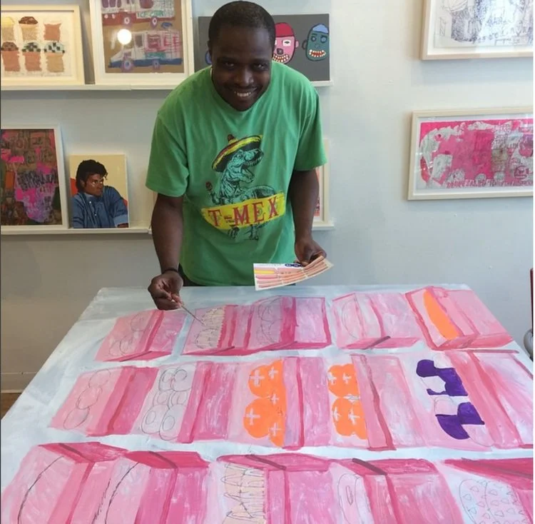 A person in a green shirt holding art supplies stands smiling near a table with colorful abstract paintings in an art studio setting, with other artworks displayed on the wall behind.