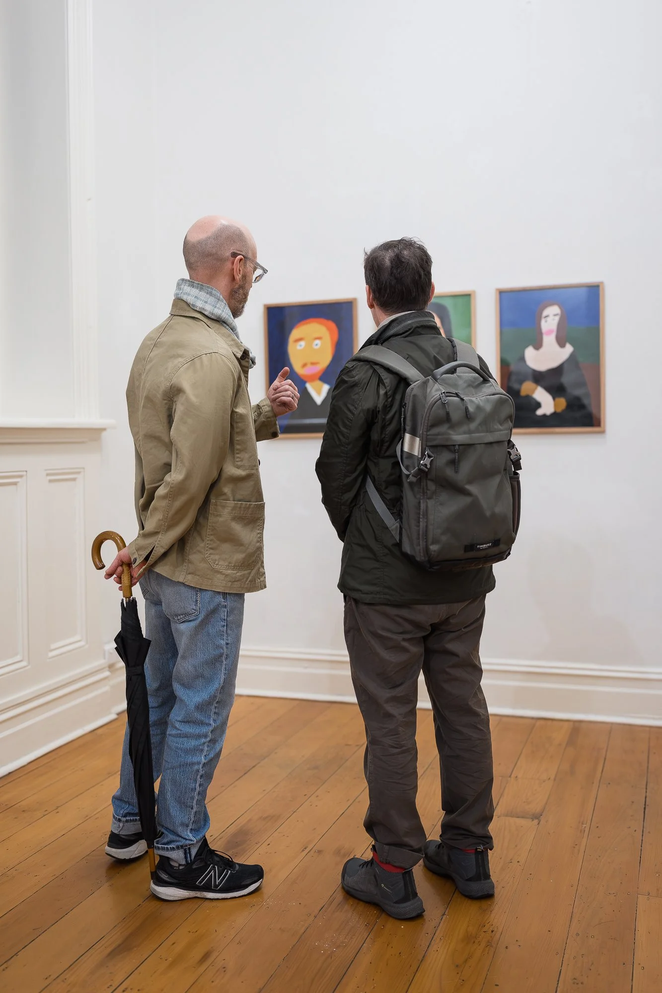 Two men viewed from behind discussing a row of three portrait paintings on a gallery wall, one gesturing with his hand.